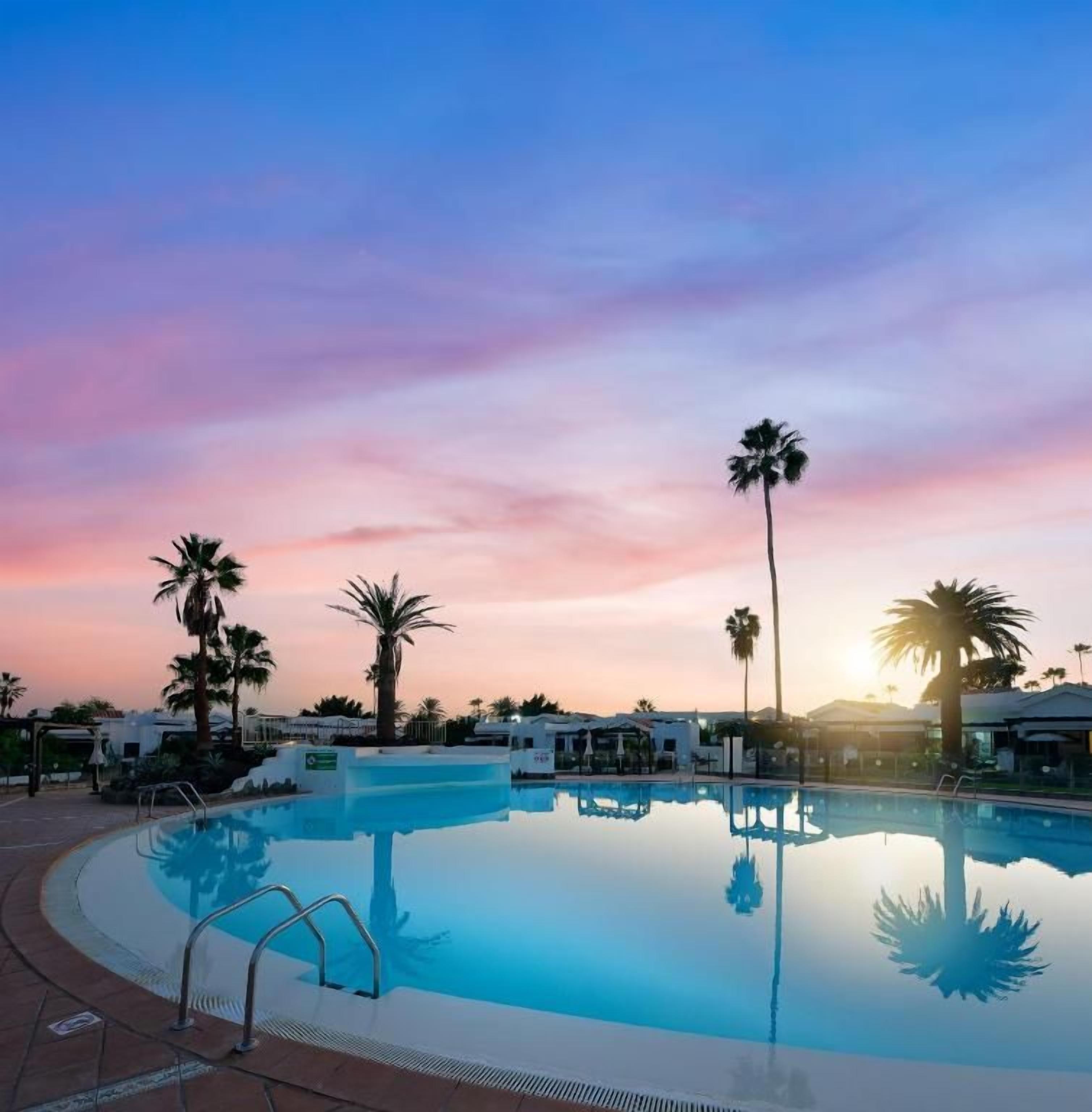Una piscina con agua cristalina, rodeada de palmeras y edificios, bajo un cielo al atardecer.