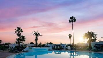 Una piscina con agua cristalina, rodeada de palmeras y edificios, bajo un cielo al atardecer.