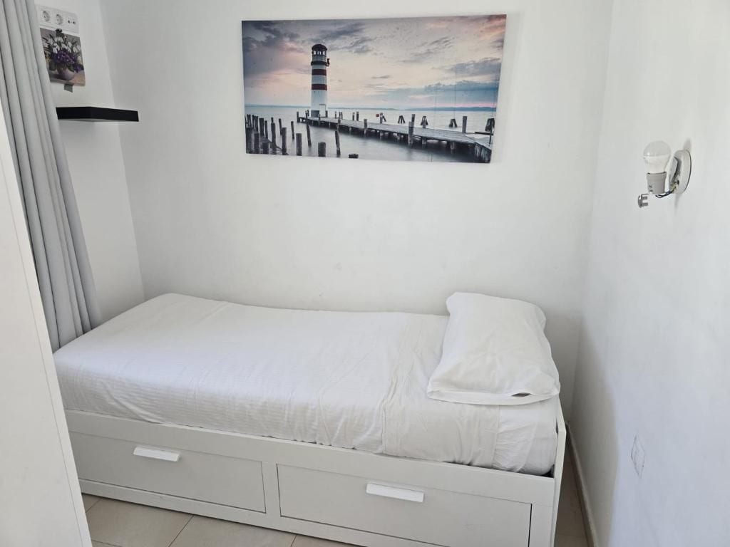 A single-bed room with a white bed, a wall-mounted picture of a lighthouse, and a small bedside table with a cup.