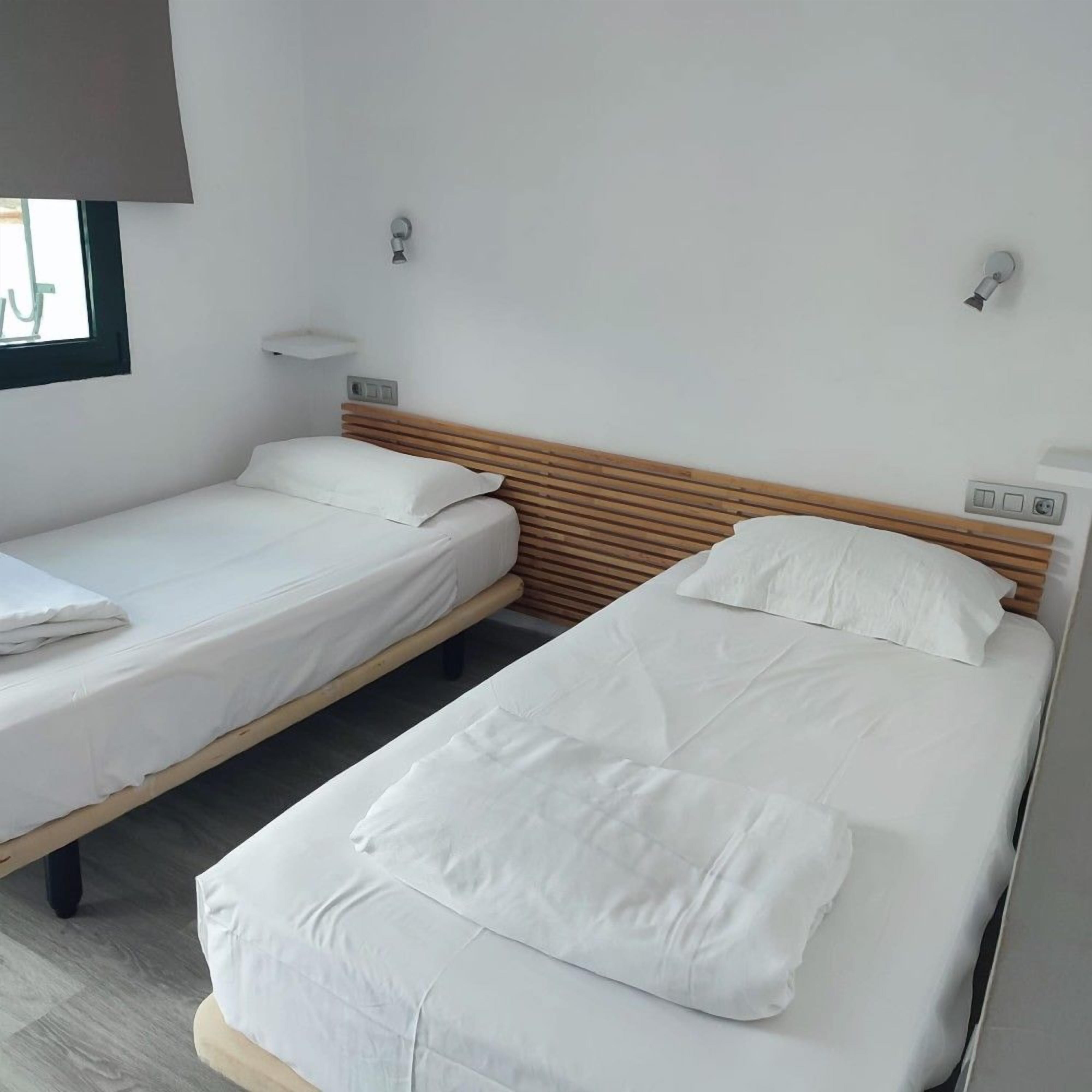 Habitación de hotel con dos camas individuales, cabecera de madera, una mesita de noche pequeña y ventana con vistas.