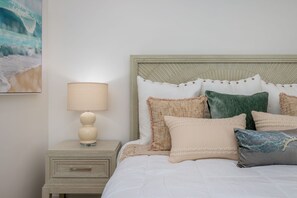 Un lit bien fait, avec une couette blanche et plusieurs oreillers, une lampe de chevet et une petite table de nuit en bois dotée d’un tiroir.