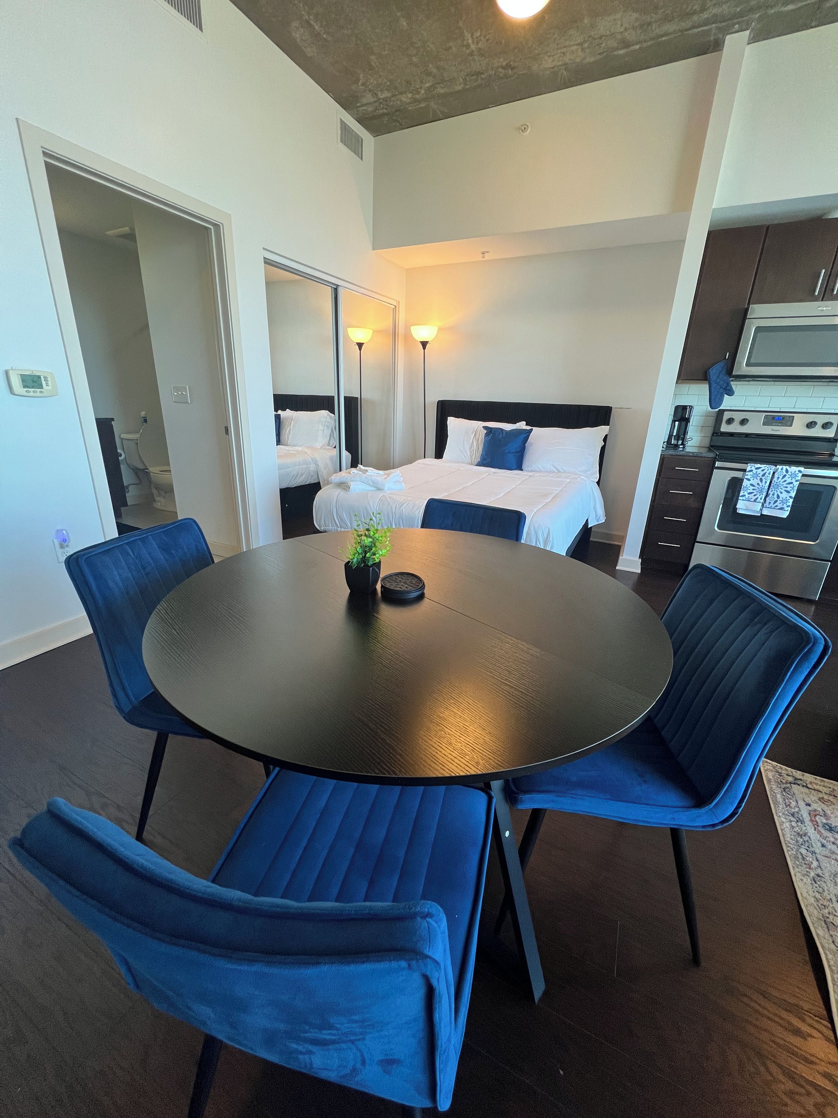 A hotel room with a round dining table, blue upholstered chairs, a bed with white bedding, a large mirror, and a kitchenette with an oven.
