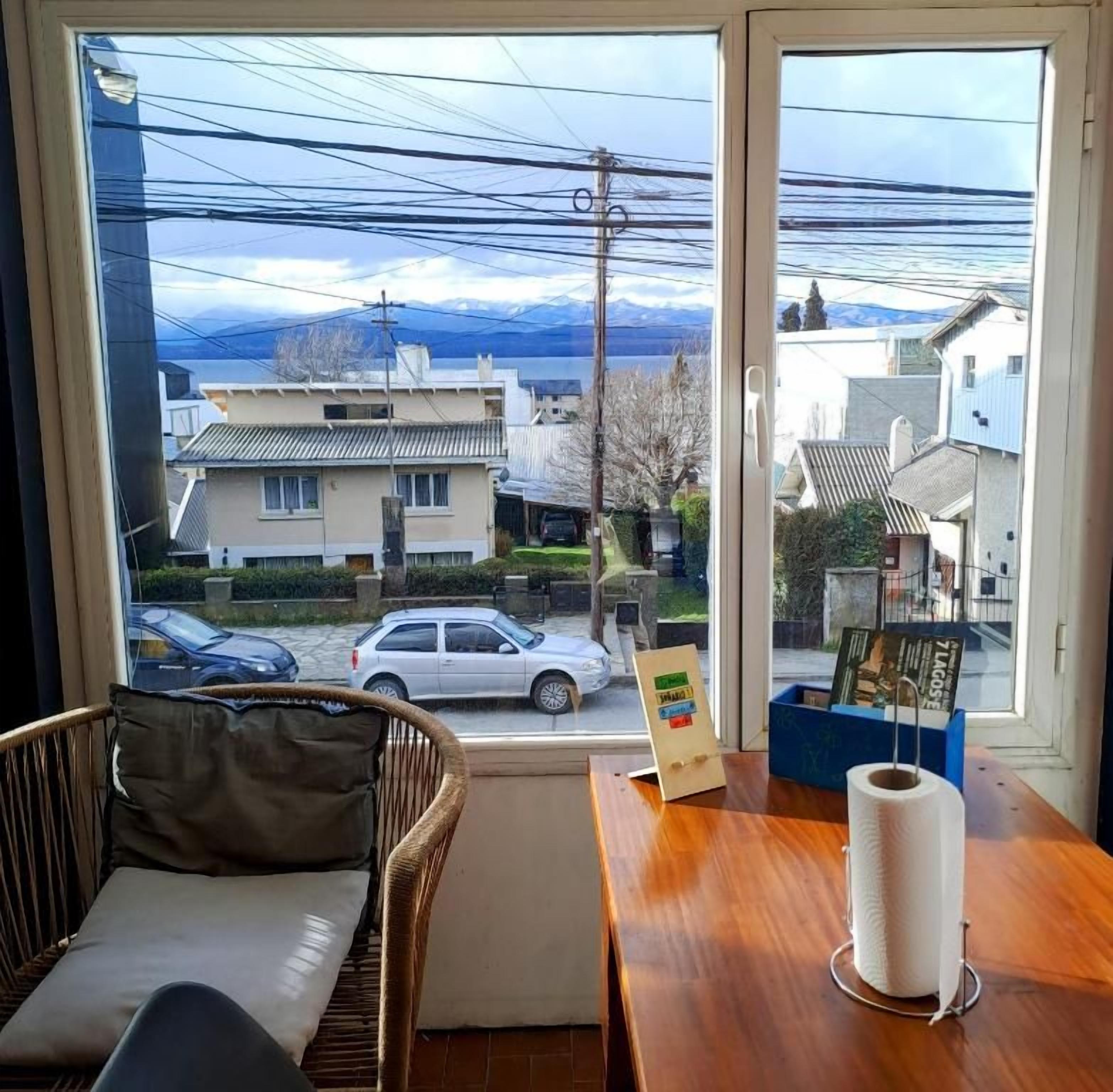 A room with a view of a residential street, a wicker chair, a wooden table with a roll of toilet paper, and a framed picture on the table.