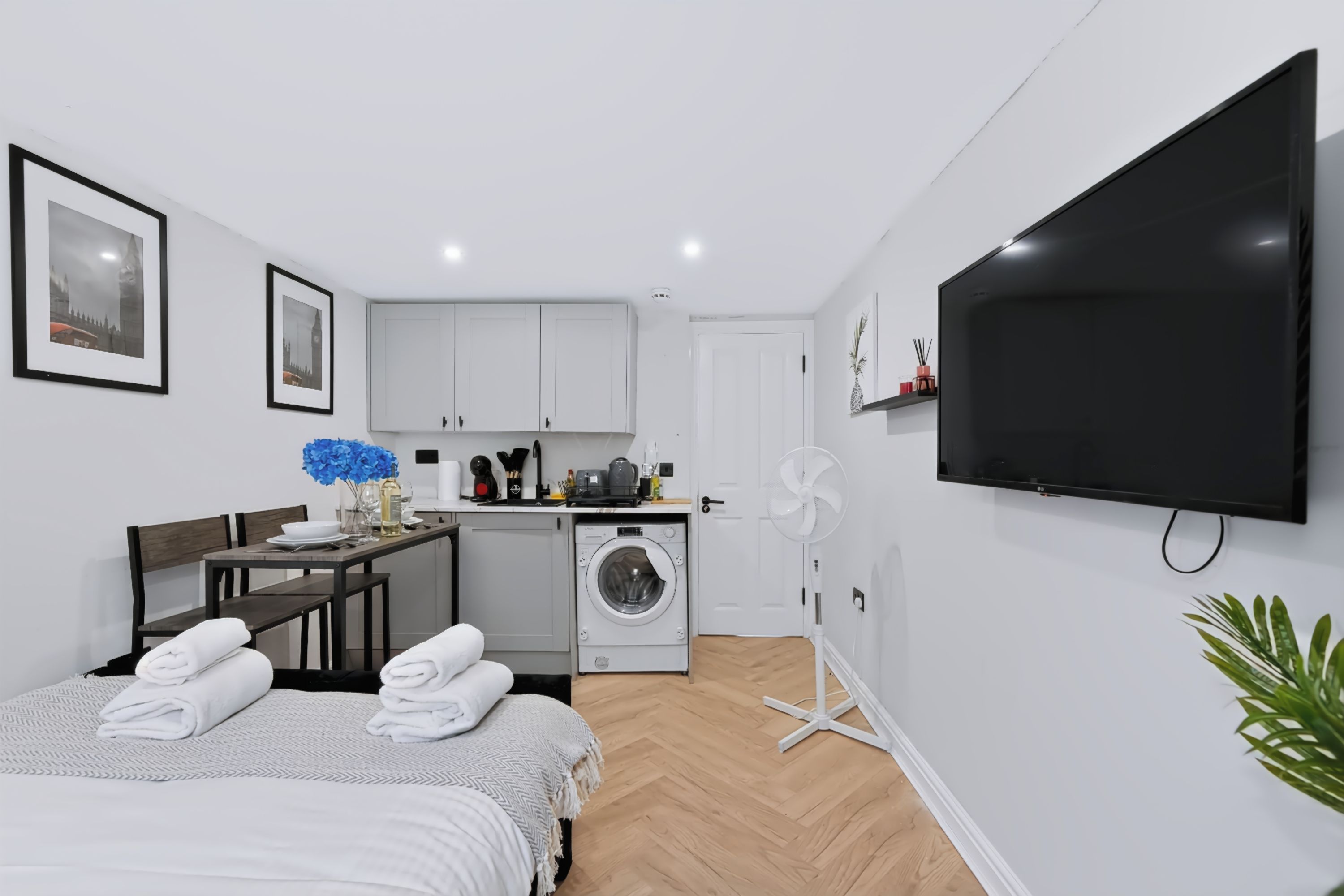 A modern hotel room with a bed, a dining area, a kitchen, a washing machine, and a TV.