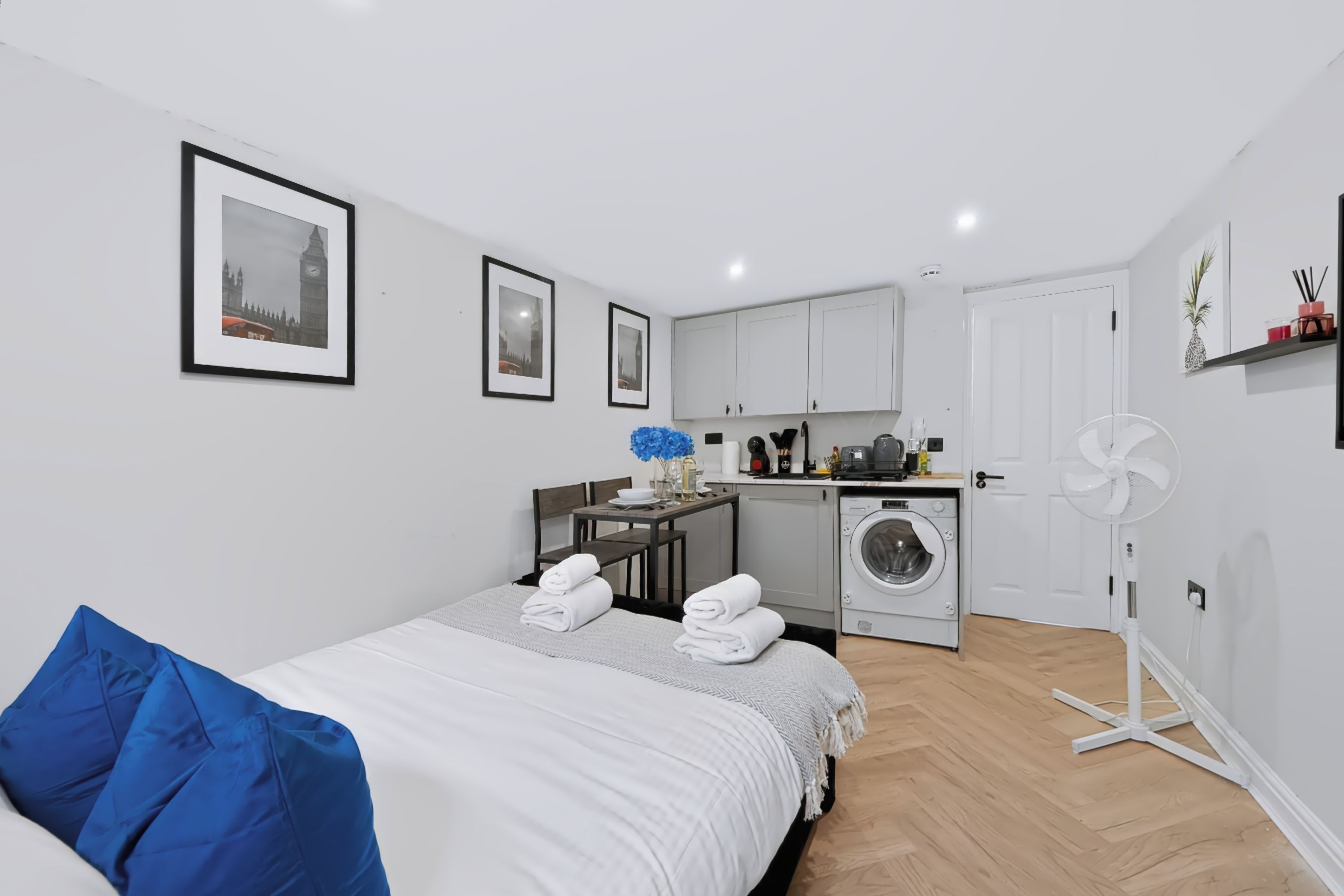Une chambre d’hôtel avec un lit, un oreiller bleu, un lave-linge, un ventilateur et des tableaux encadrés accrochés au mur.