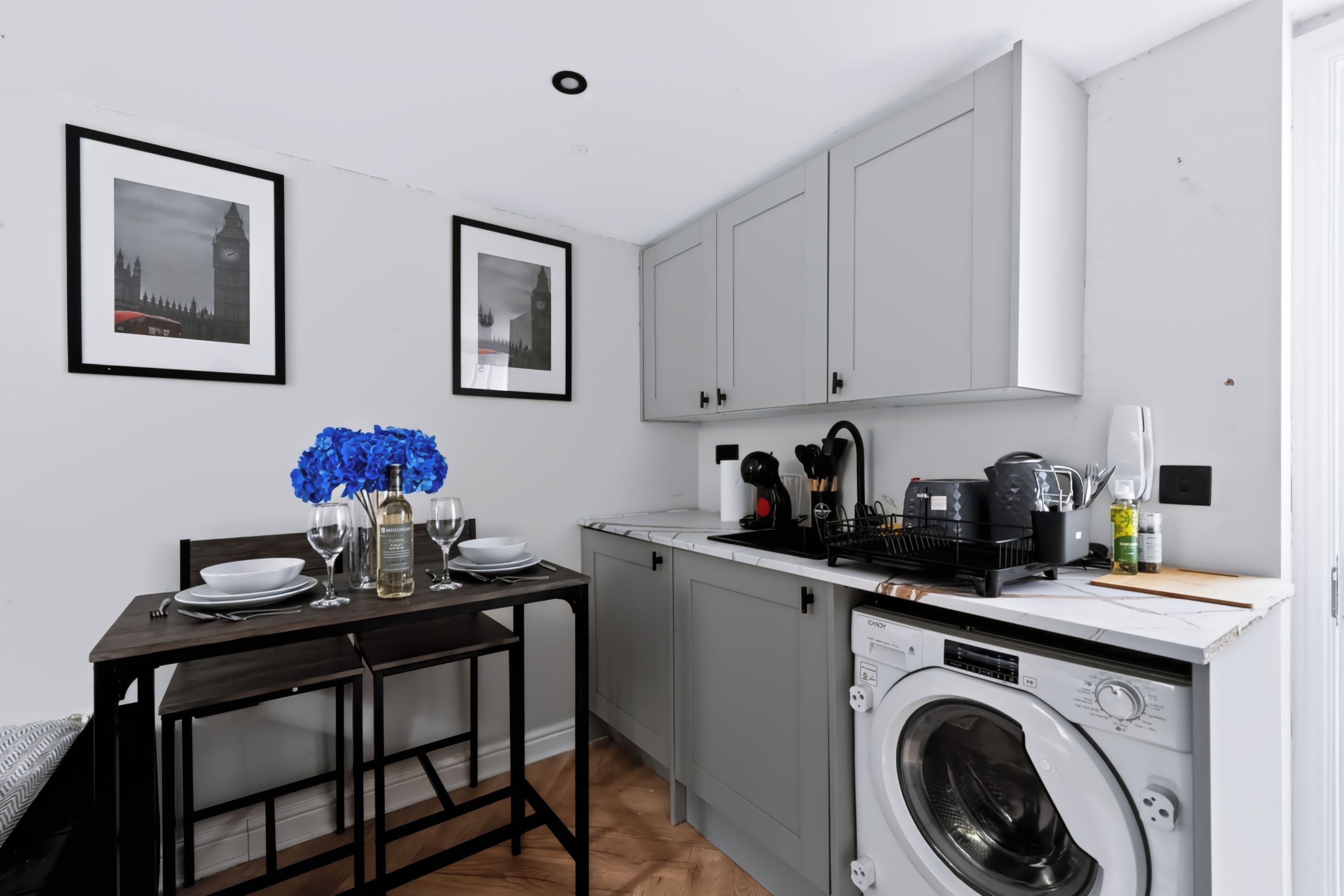 Une cuisine compacte équipée d’un lave-linge intégré, d’une table à manger pour deux personnes et de tableaux encadrés accrochés au mur.