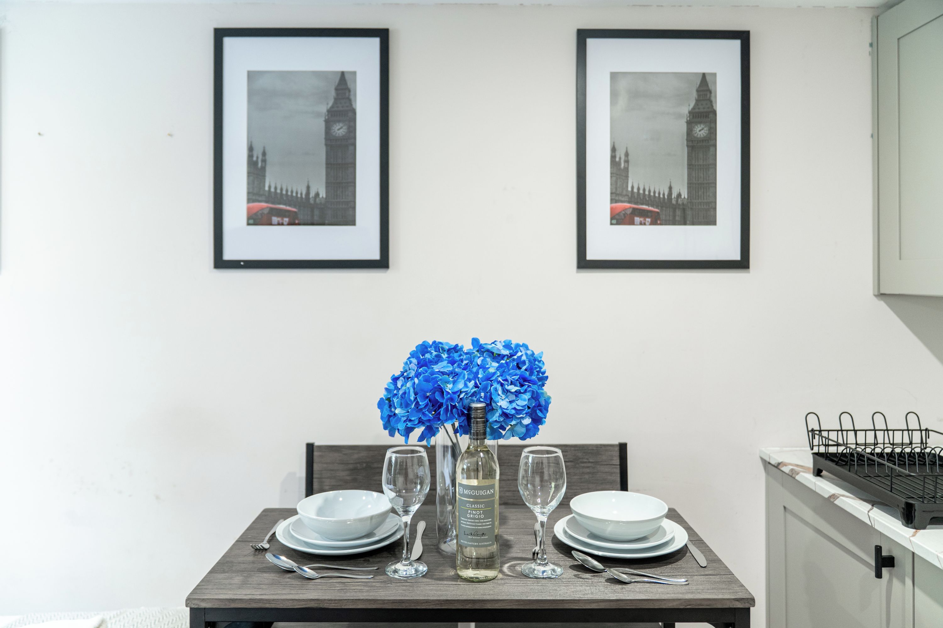 A dining table with plates, glasses, cutlery, and a bottle of wine, flanked by two framed pictures of a clock tower.