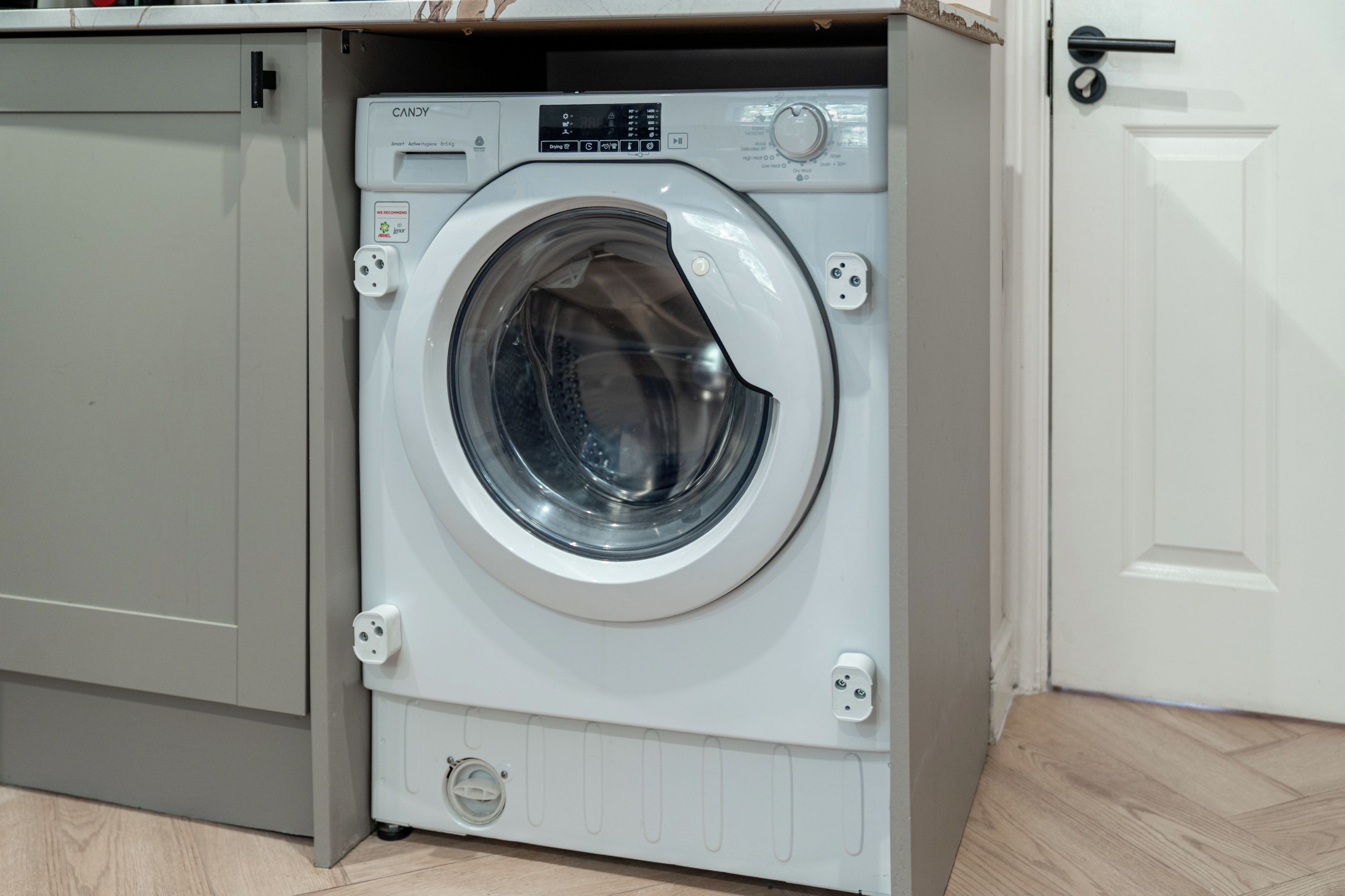 Un lave-linge à chargement frontal doté d’un panneau de commande, situé dans une pièce dont le sol est en bois et la porte est blanche.