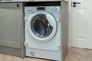 A front-loading washing machine with a control panel, located in a room with a wooden floor and a white door. - Budget studio apartments - Streatham SW16 (London)