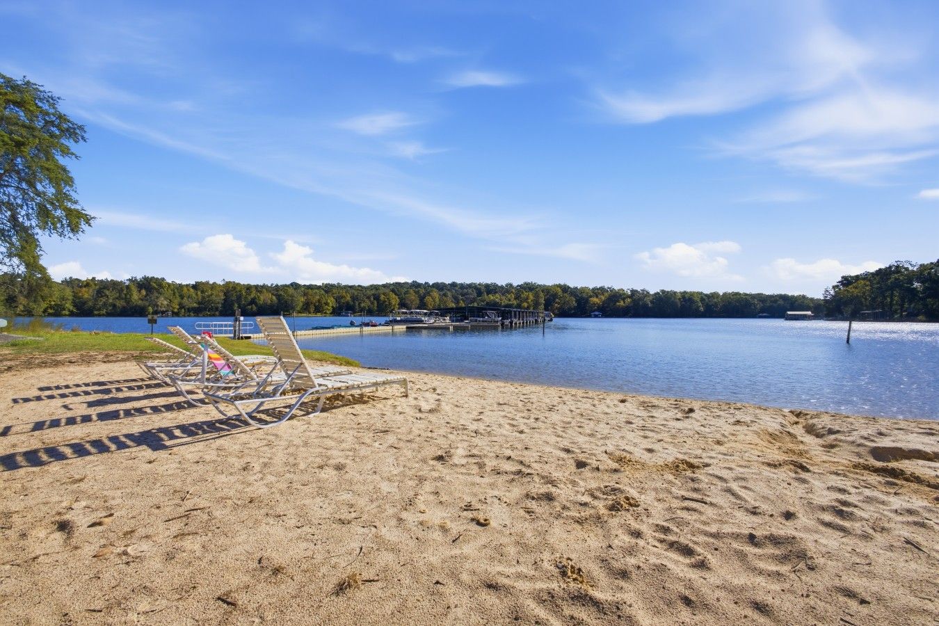 A sandy beach with lounge chairs and a dock extending into a calm lake, surrounded by trees.