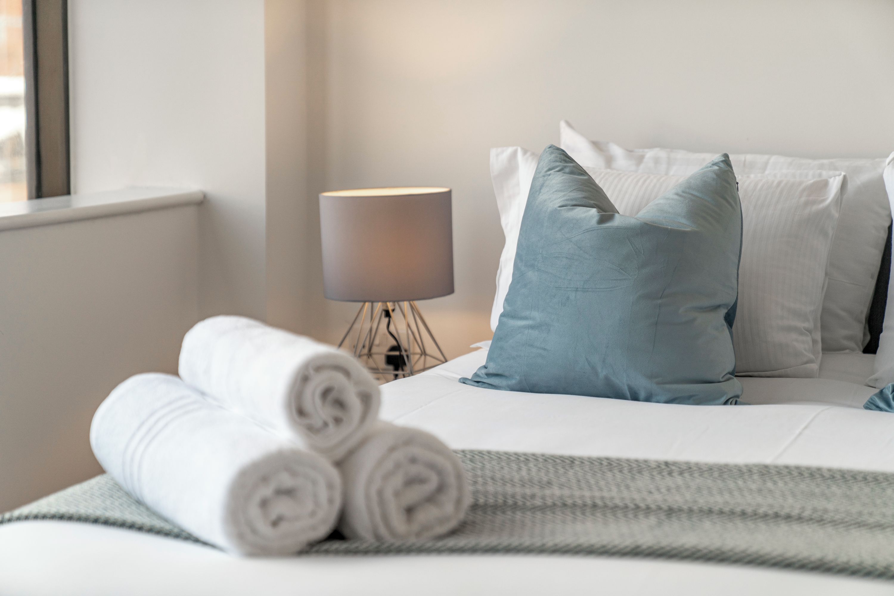 A neatly made bed with white linens and rolled towels, a grey throw blanket, and a lamp on a bedside table.