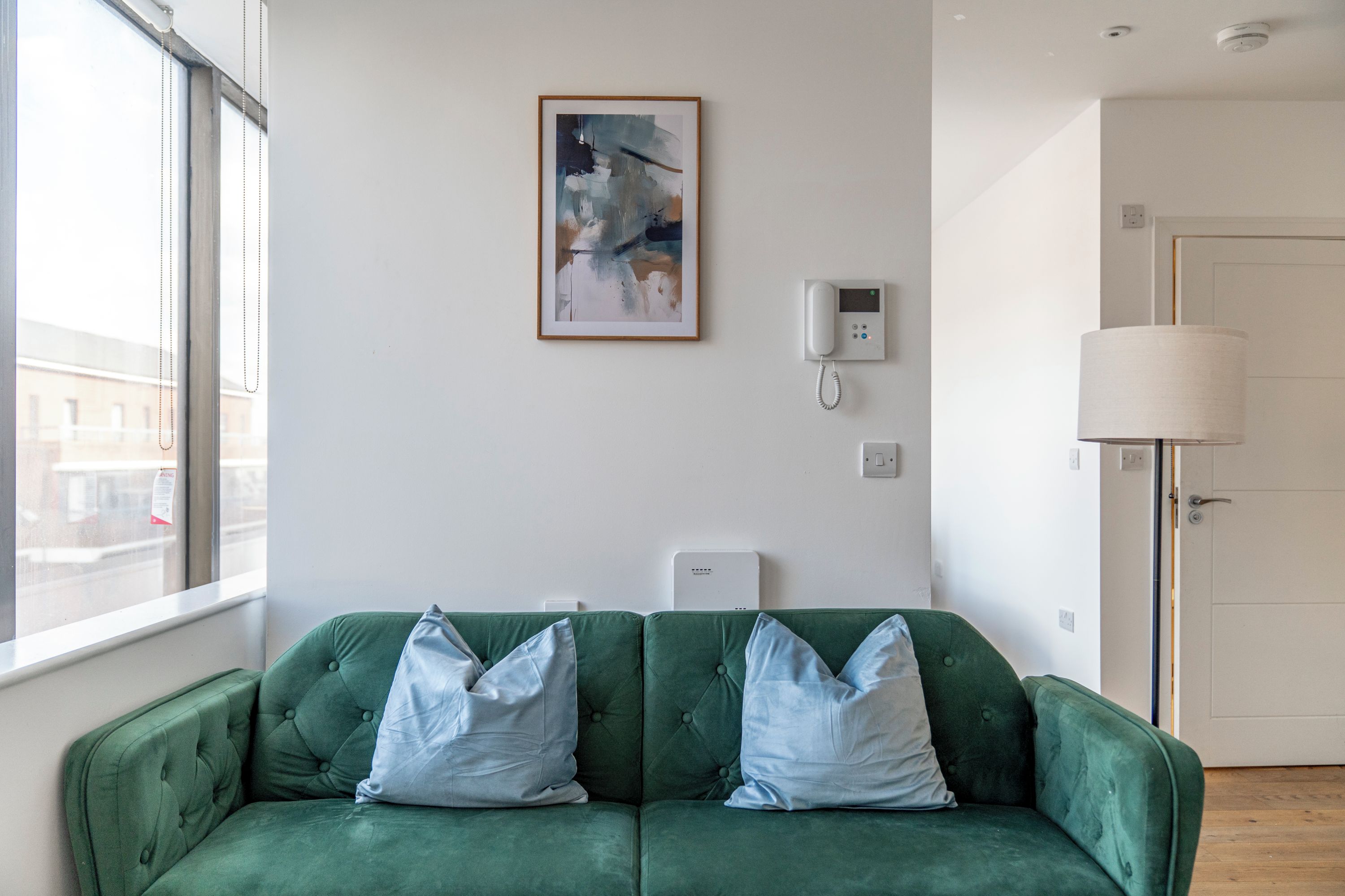 A green sofa with two pillows, a wall-mounted phone, a framed abstract painting, and a lamp.
