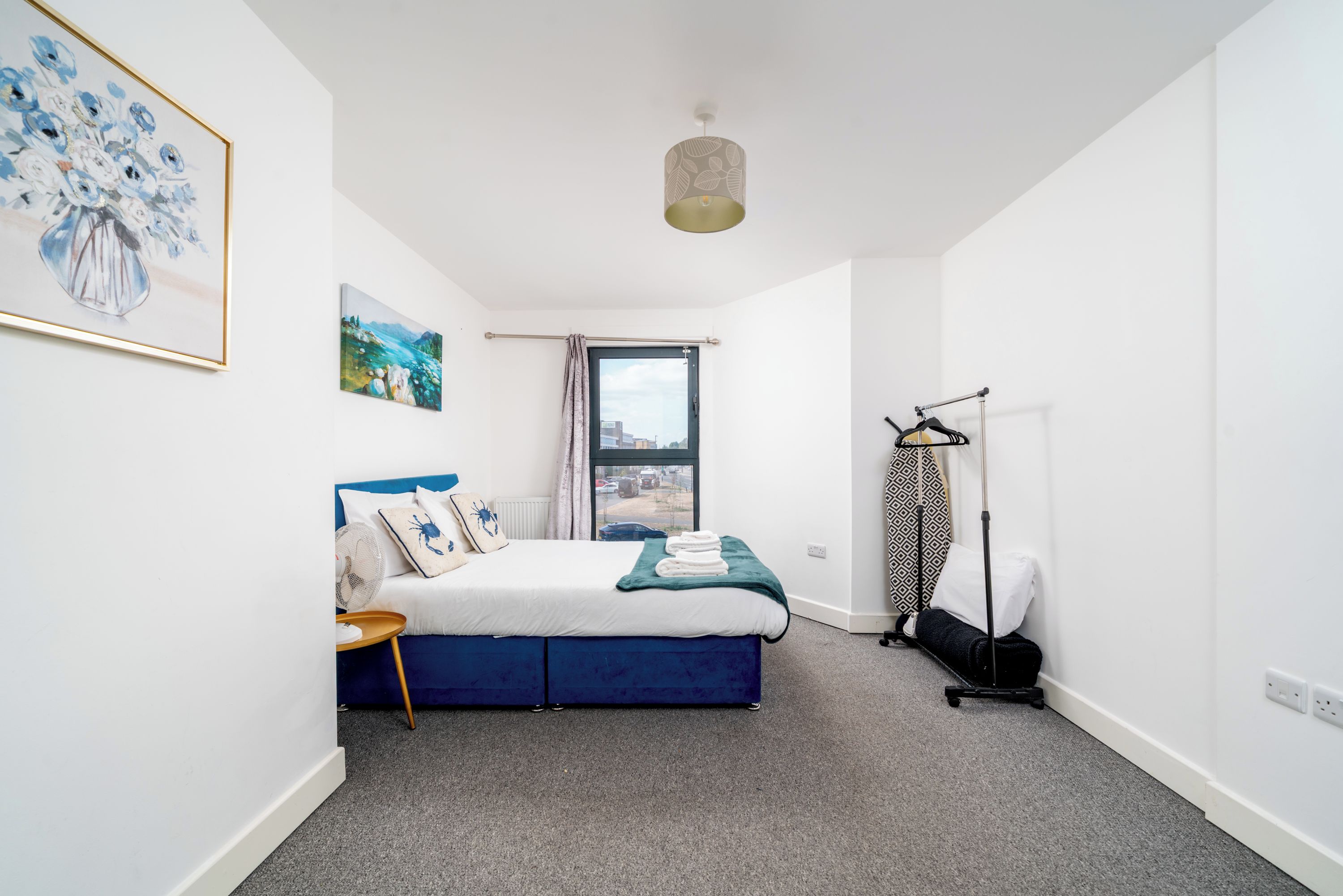 Een slaapkamer met een bed, raam, radiator, ventilator en een kledingrek.