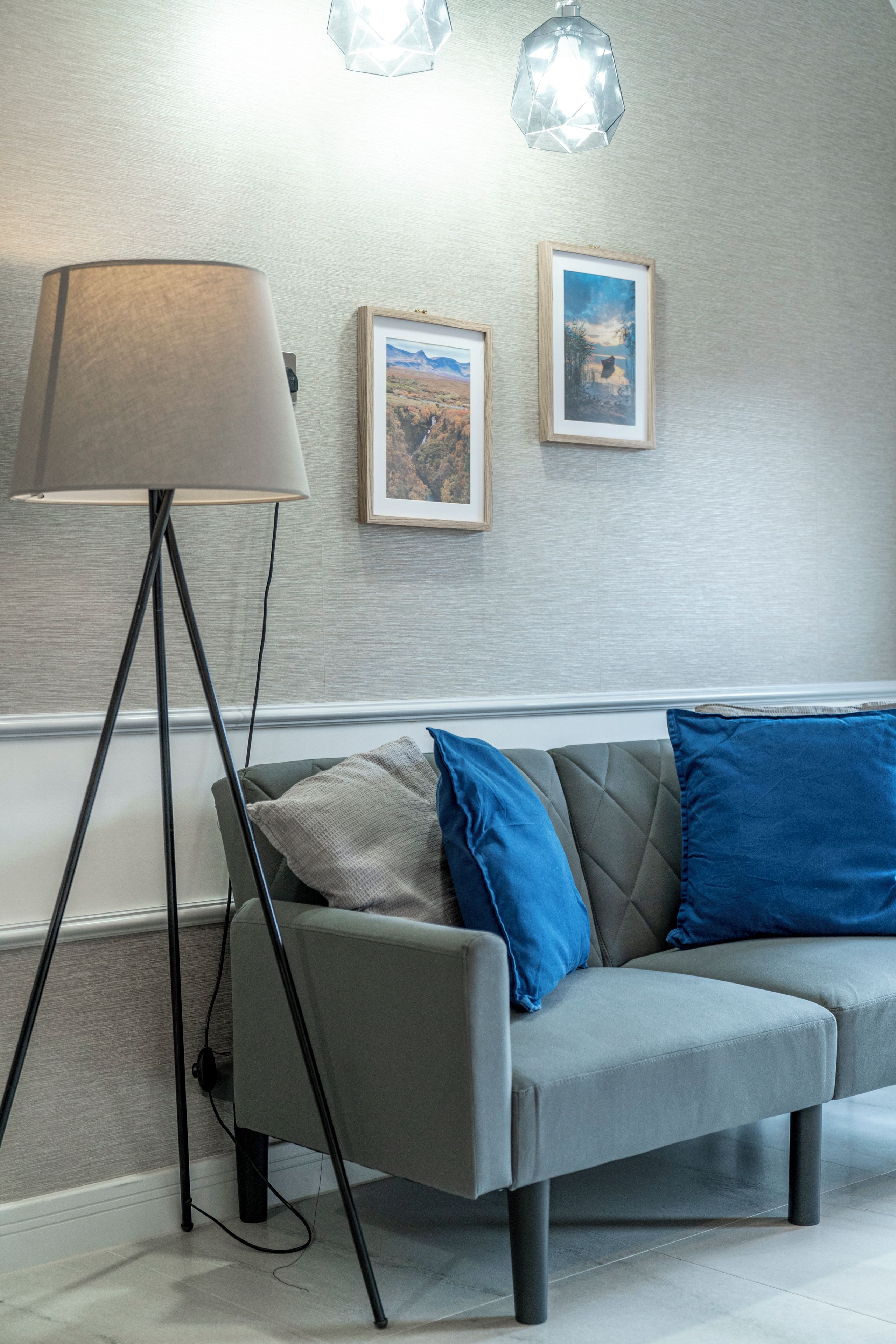 Ein modernes Wohnzimmer mit einem grauen Sofa, zwei blauen Kissen, einer Stehlampe und zwei gerahmten Landschaftsbildern an der Wand.