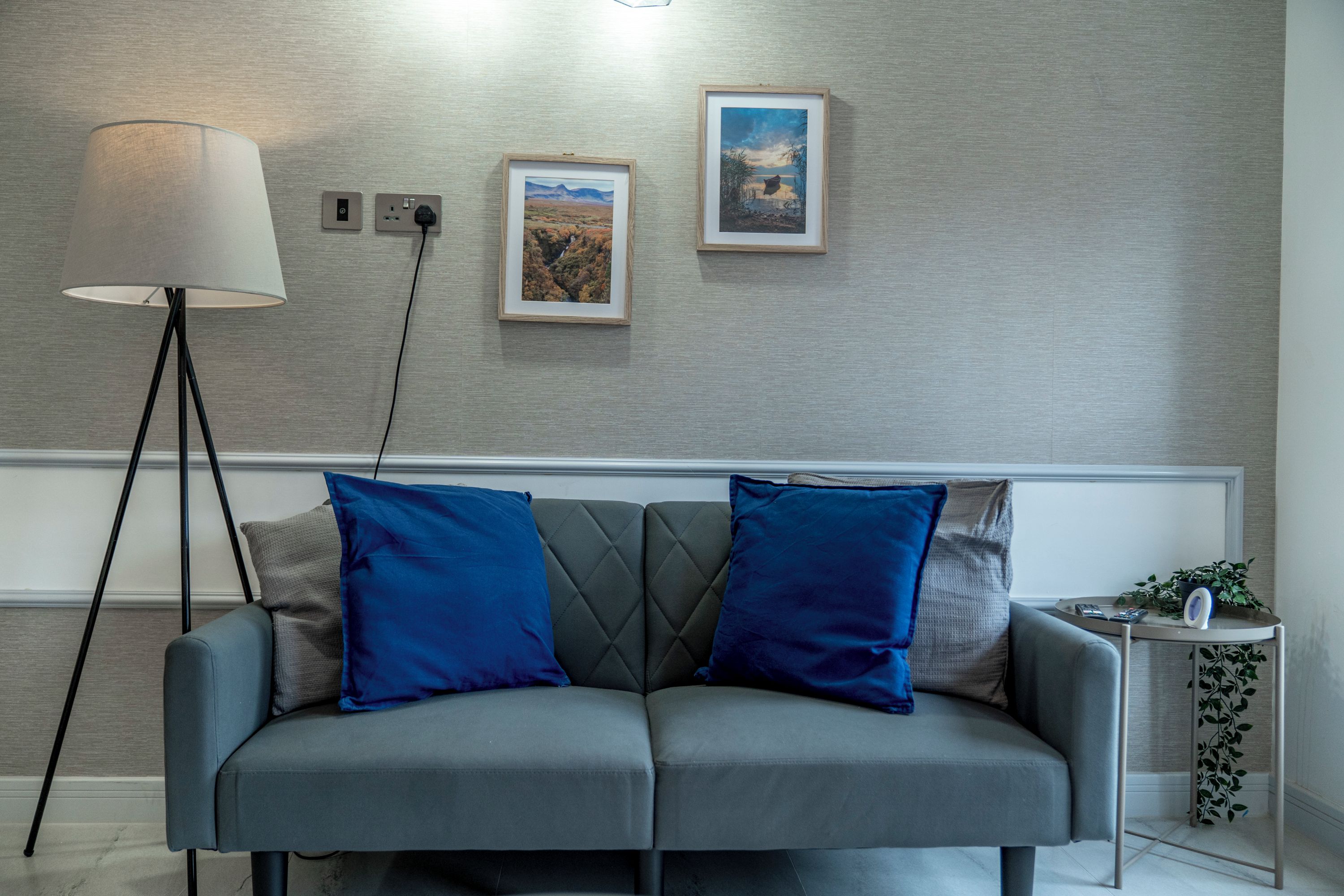Ett modernt vardagsrum med en grå soffa, två blå kuddar, en golvlampa och två inramade tavlor på väggen.