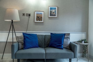Ett modernt vardagsrum med en grå soffa, två blå kuddar, en golvlampa och två inramade tavlor på väggen.