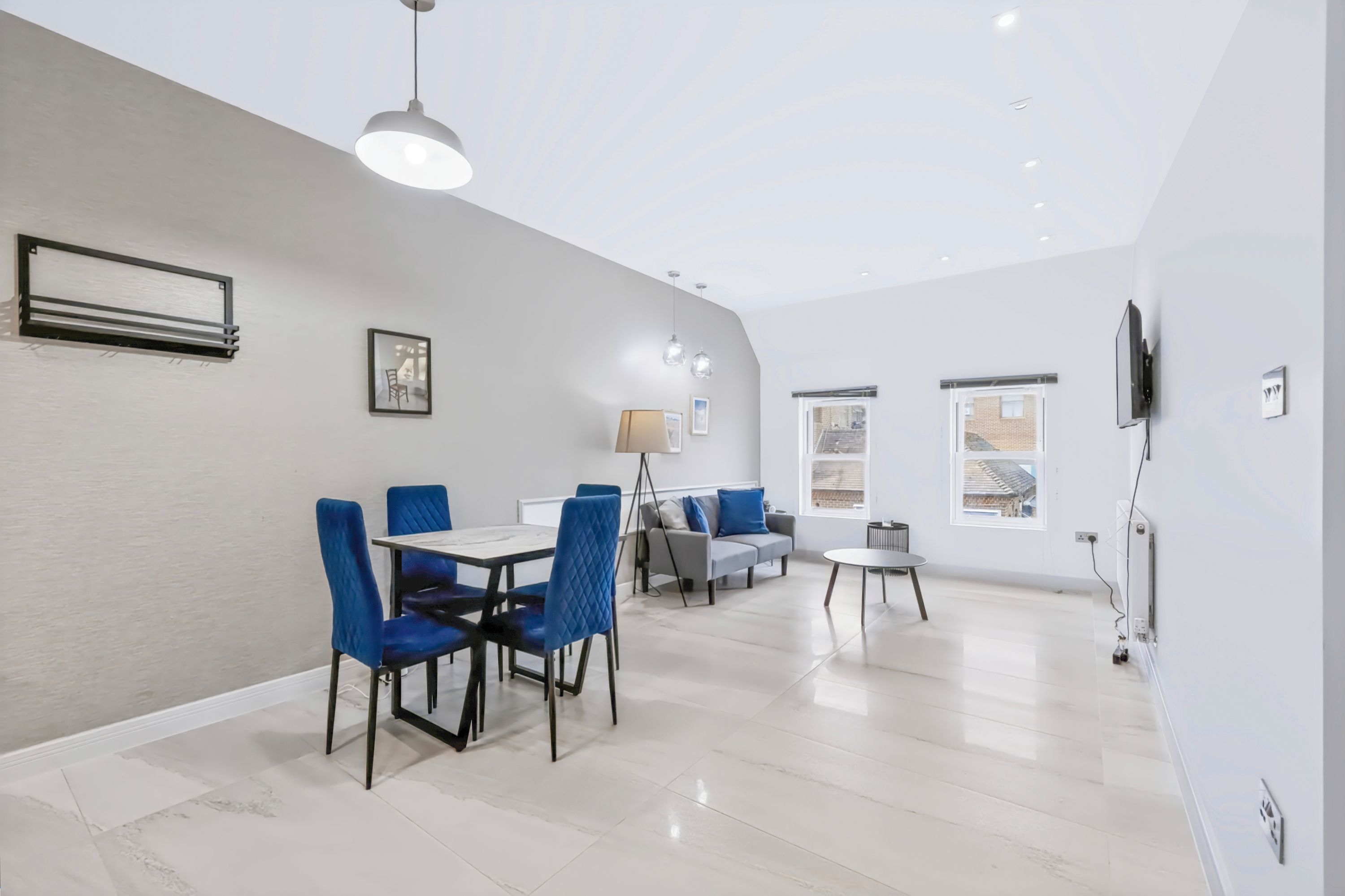 A modern living room with a dining area, a sofa, a TV mounted on the wall, and a large window.
