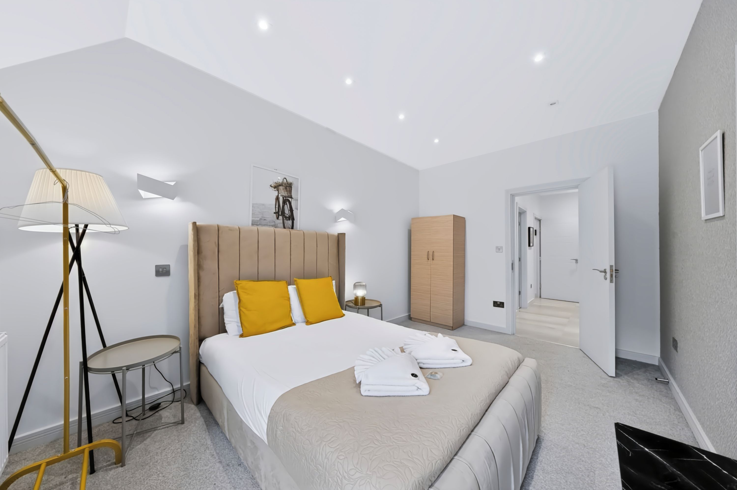A modern hotel room with a large bed, a bedside table, a lamp, a wardrobe, and a door leading to another room.