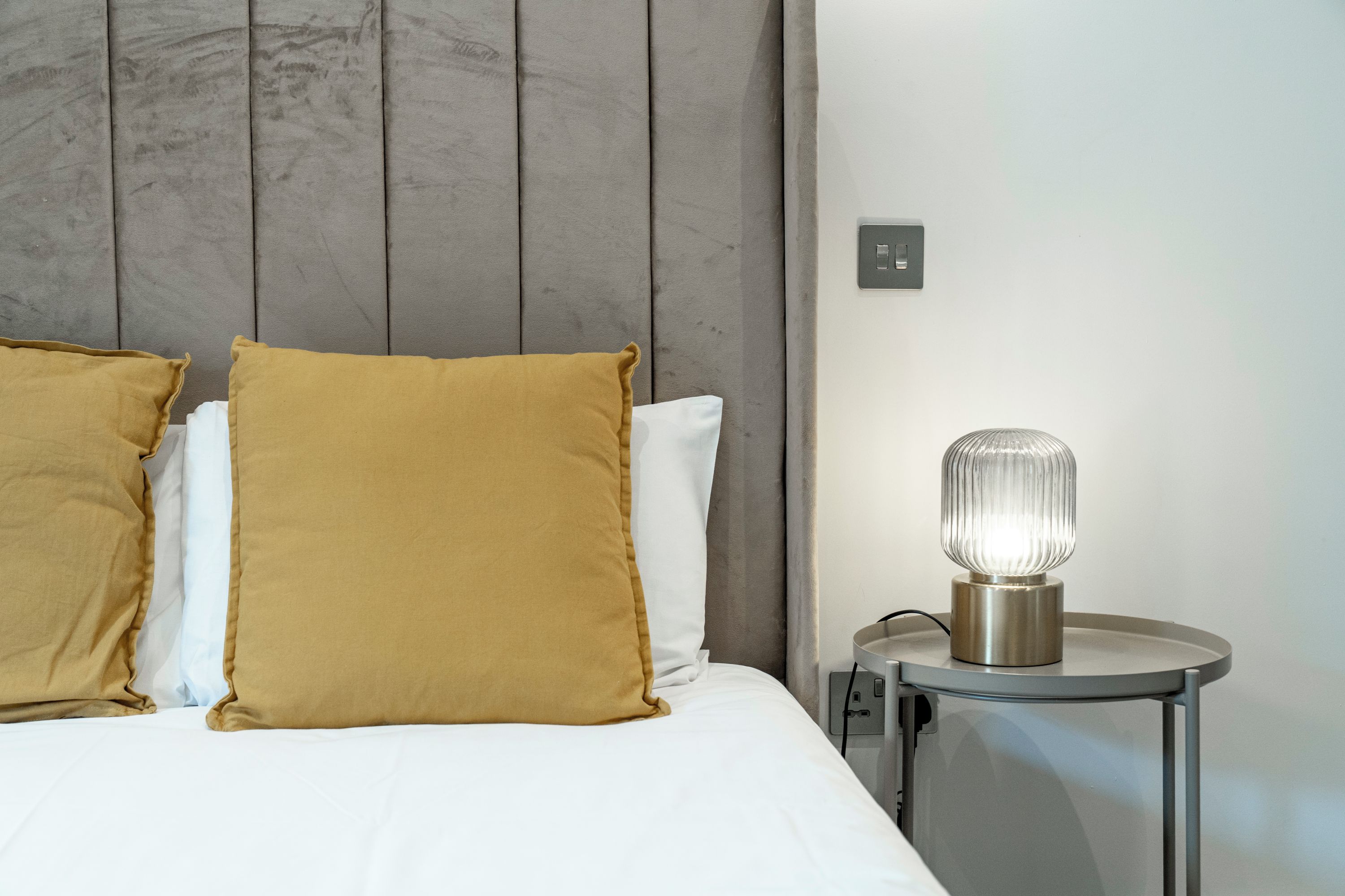 Una cama con almohadillas blancas y mostaza, un cabecero gris y una mesita de noche con una lámpara.