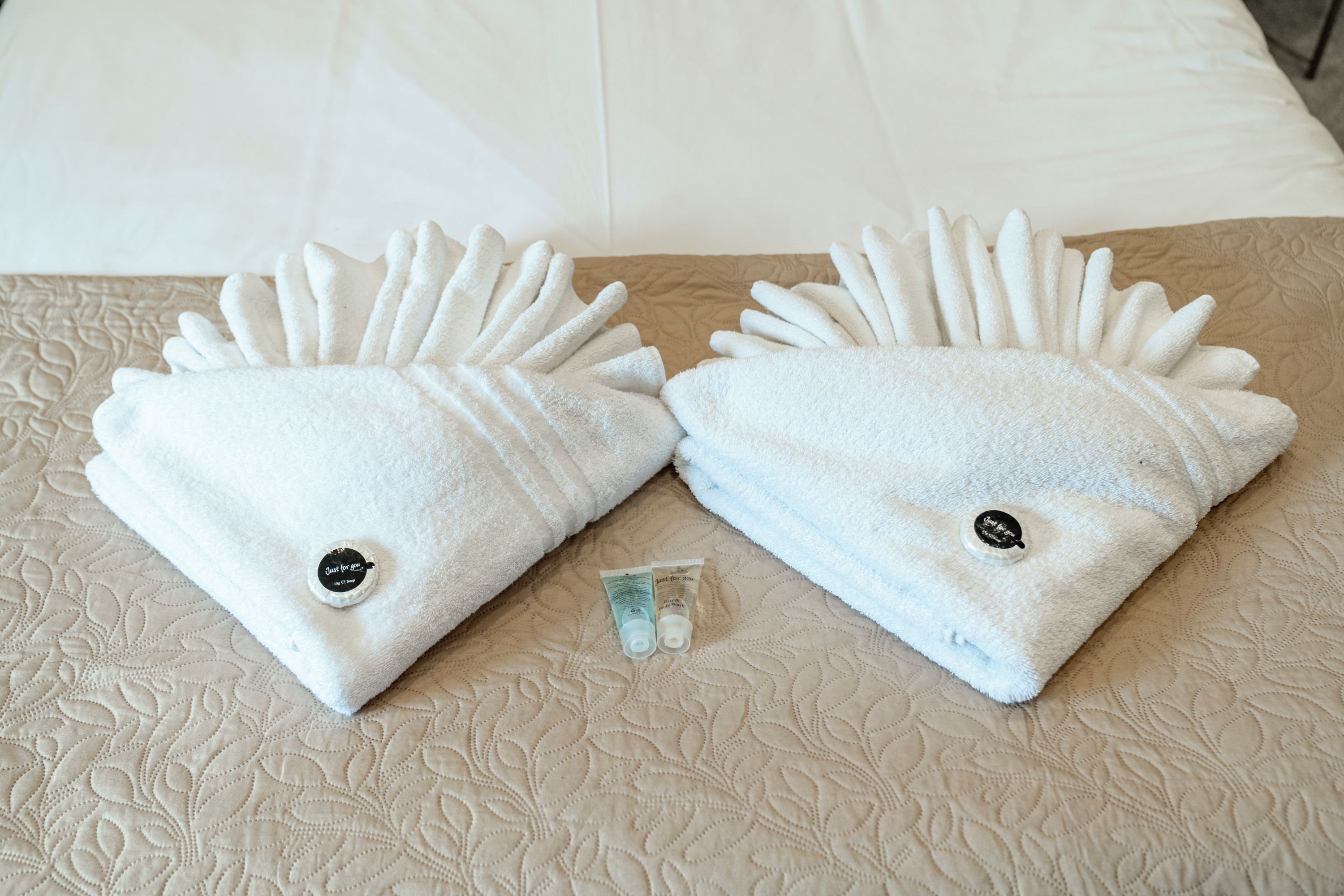 Dos toallas blancas con un botón, sobre una cama con una colcha beige texturizada.