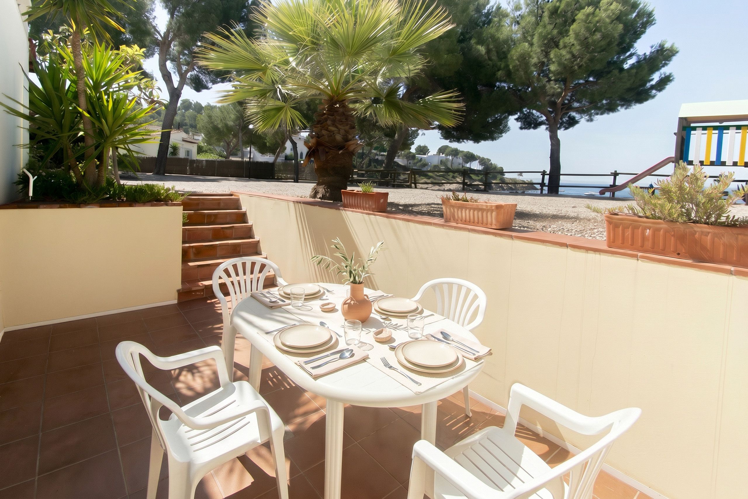 An outdoor patio with a table set for four, surrounded by white chairs, with a palm tree and a view of the sea in the background.