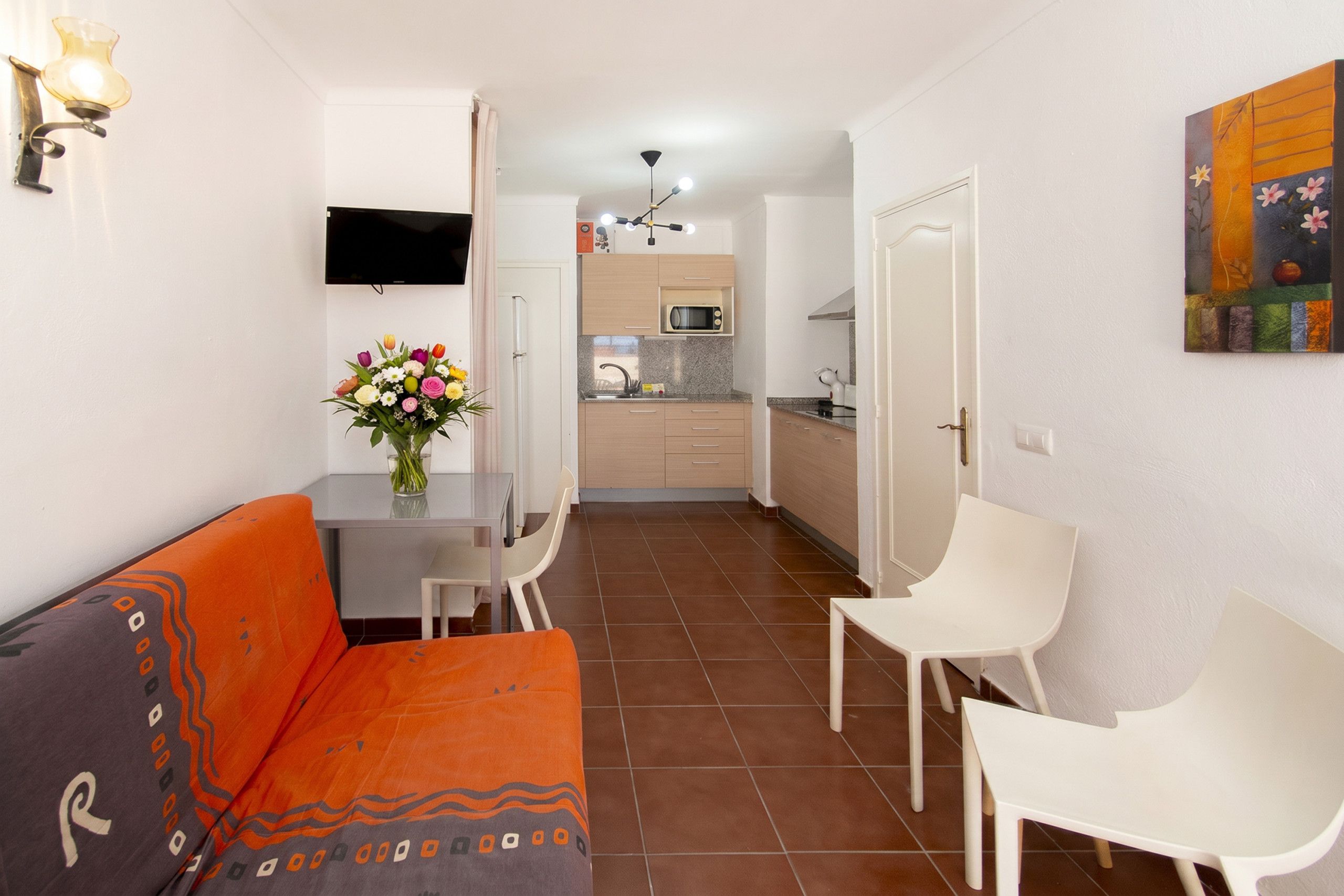A compact living space with a sofa, dining table, chairs, a kitchen area, and a TV mounted on the wall.