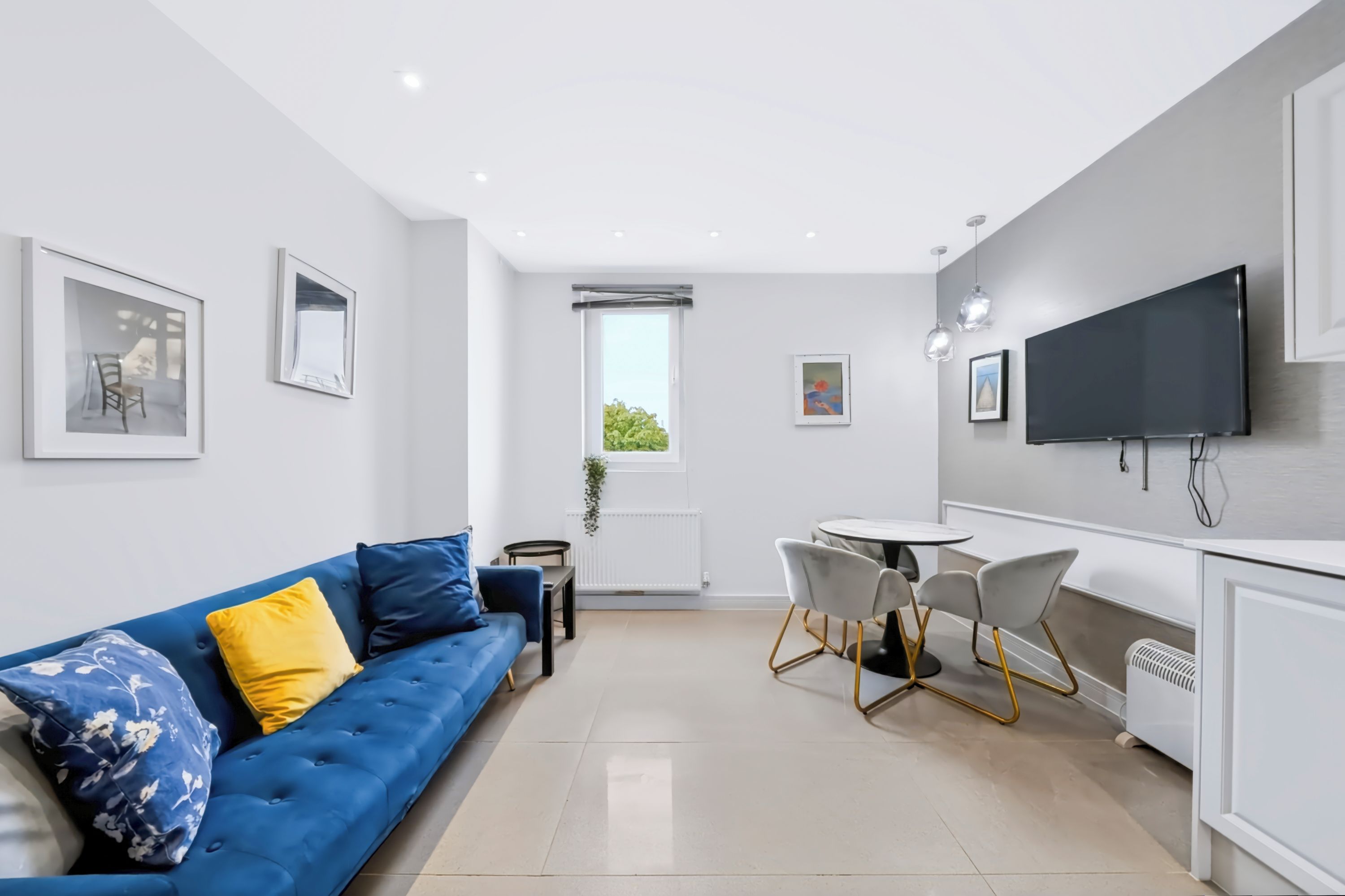 A modern living room with a blue sofa, a dining table with chairs, a flat-screen TV, and artwork on the walls.