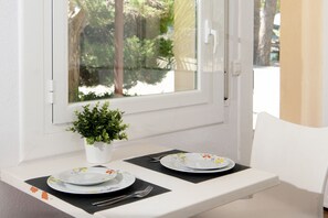 Een kleine tafel met twee borden en bestek, een bloempot en een raam met uitzicht op bomen en een hek.