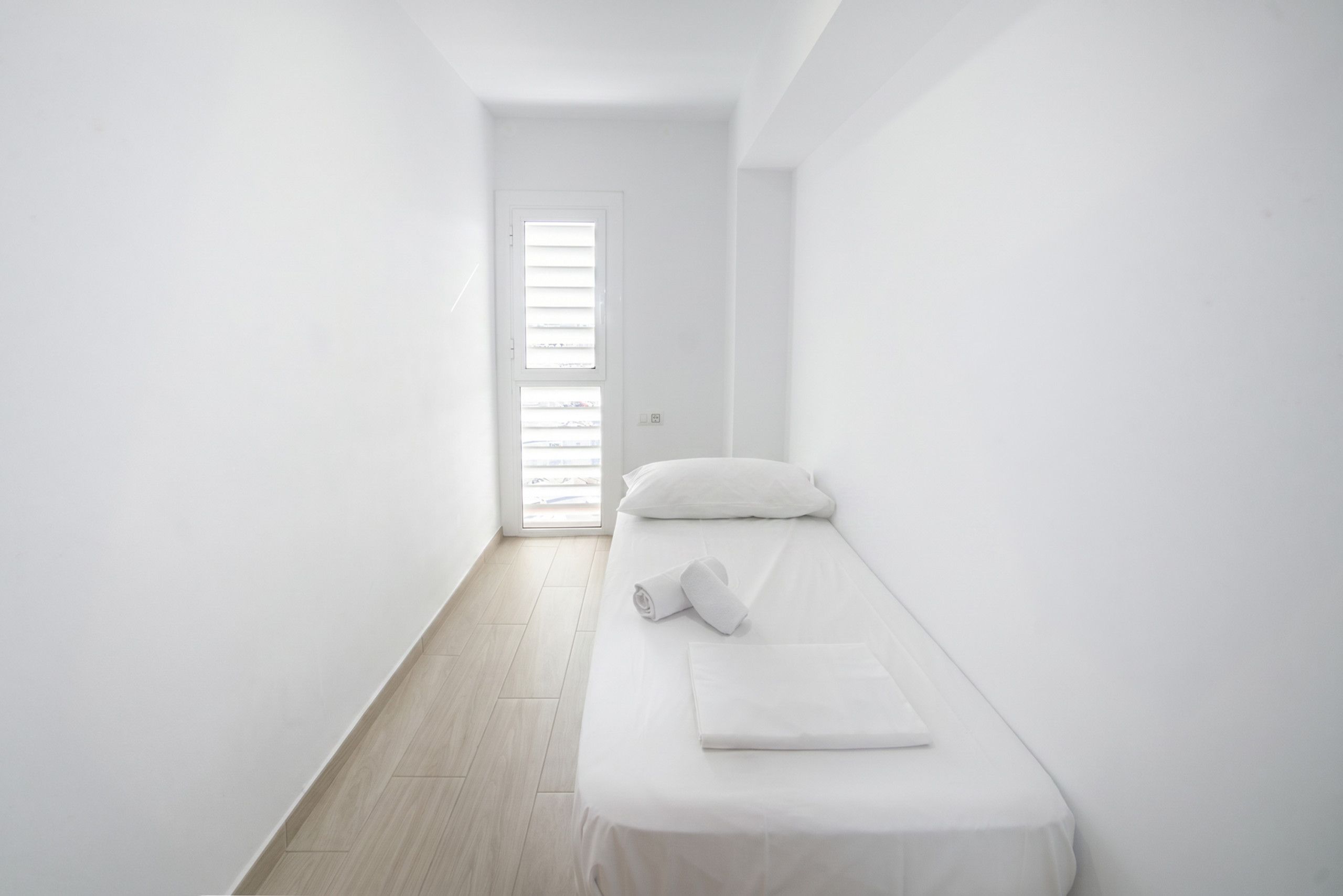 Ein schlicht eingerichtetes Zimmer mit einem Einzelbett, einer Tür mit Jalousien und einem hellen Holzboden.