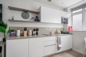 A modern kitchen with white cabinetry, a built-in microwave, a kettle, and various kitchen utensils. - Modern studio apt in West Hampstead London (London)