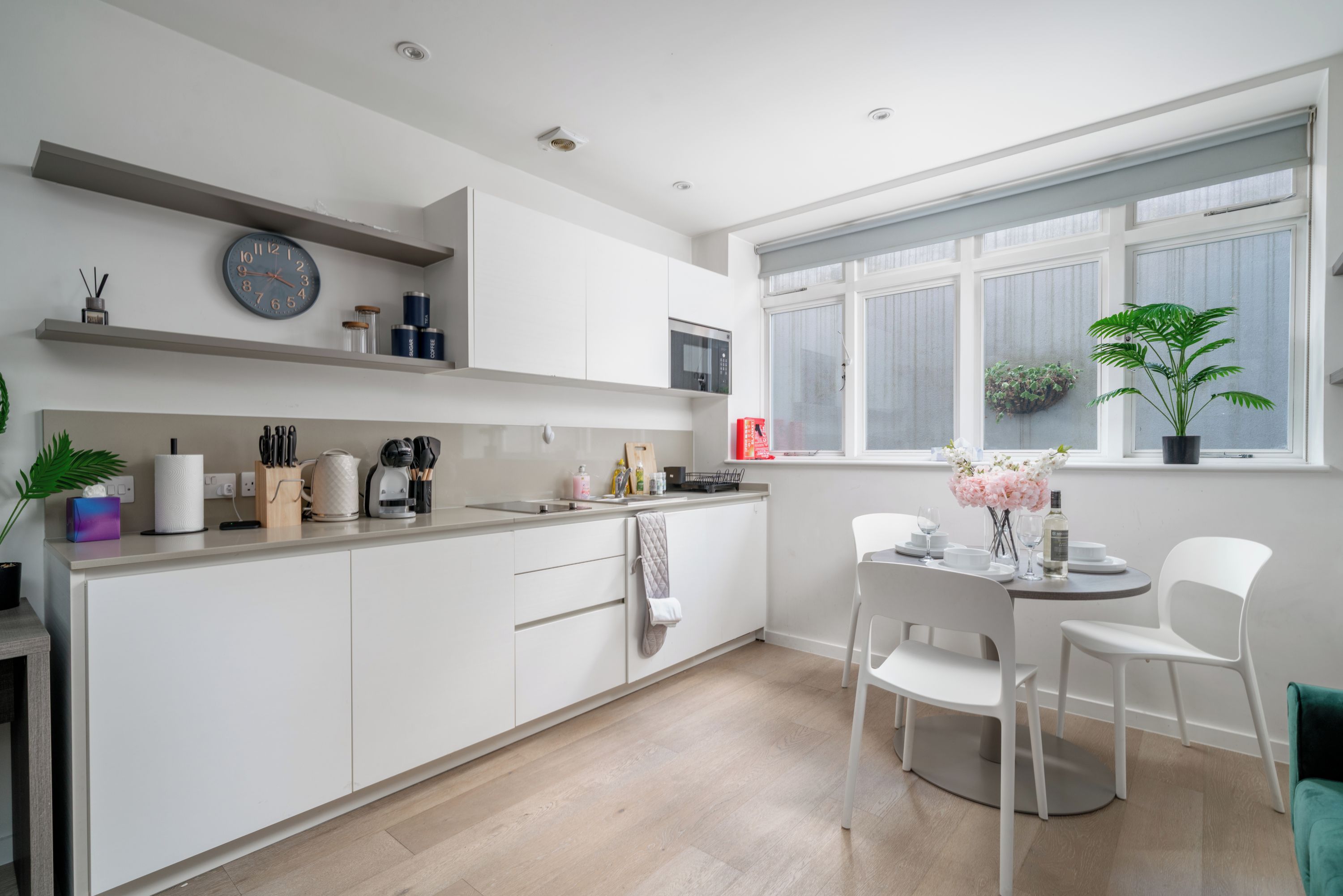 A modern kitchen with white cabinetry, a dining area with white chairs, a window with plants, and a clock on the wall.
