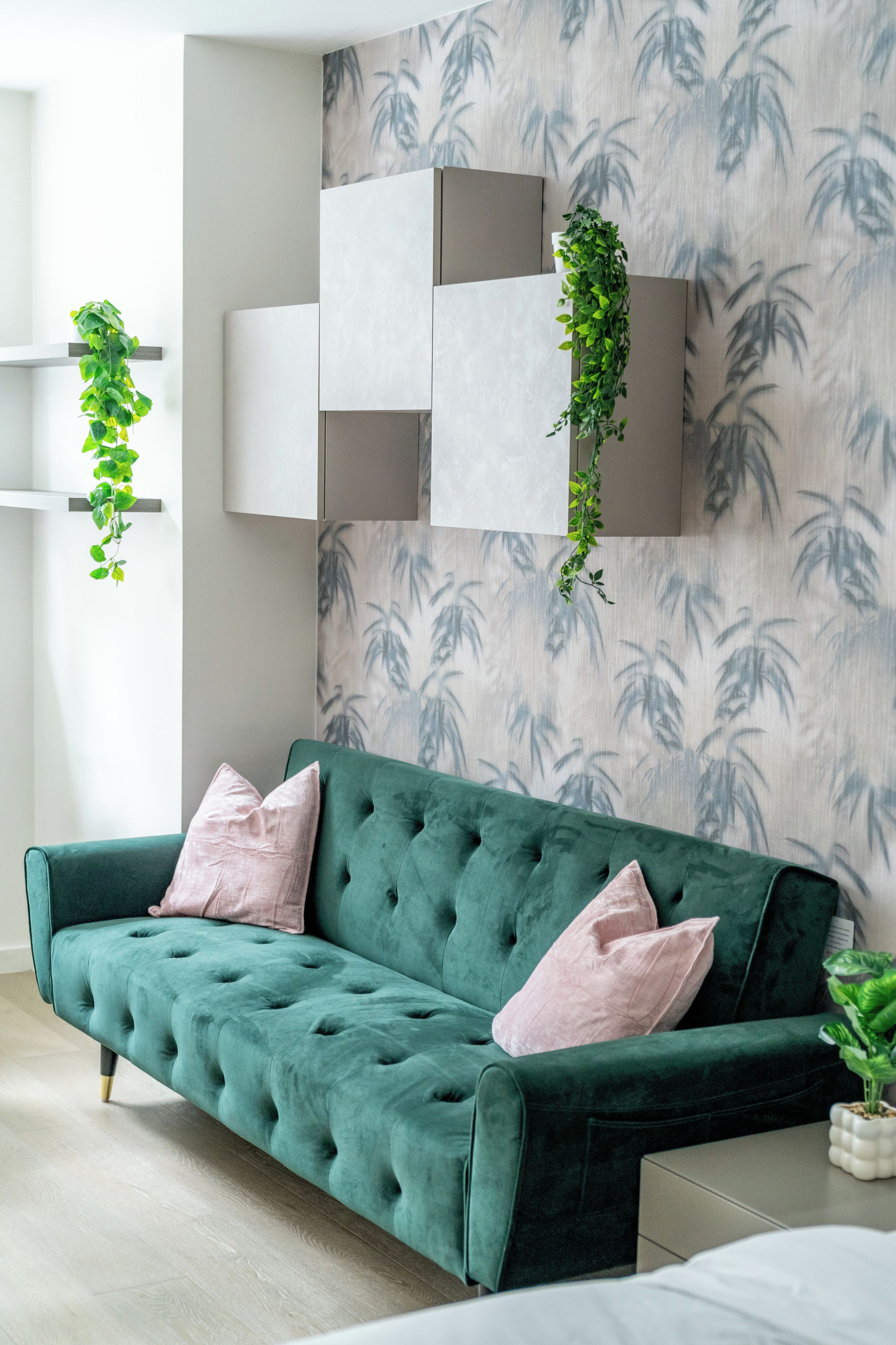 A green sofa with tufted detailing and two pink cushions, positioned against a wall with a bamboo leaf pattern.
