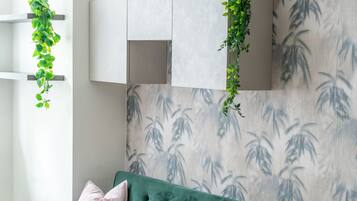 A green sofa with tufted detailing and two pink cushions, positioned against a wall with a bamboo leaf pattern.