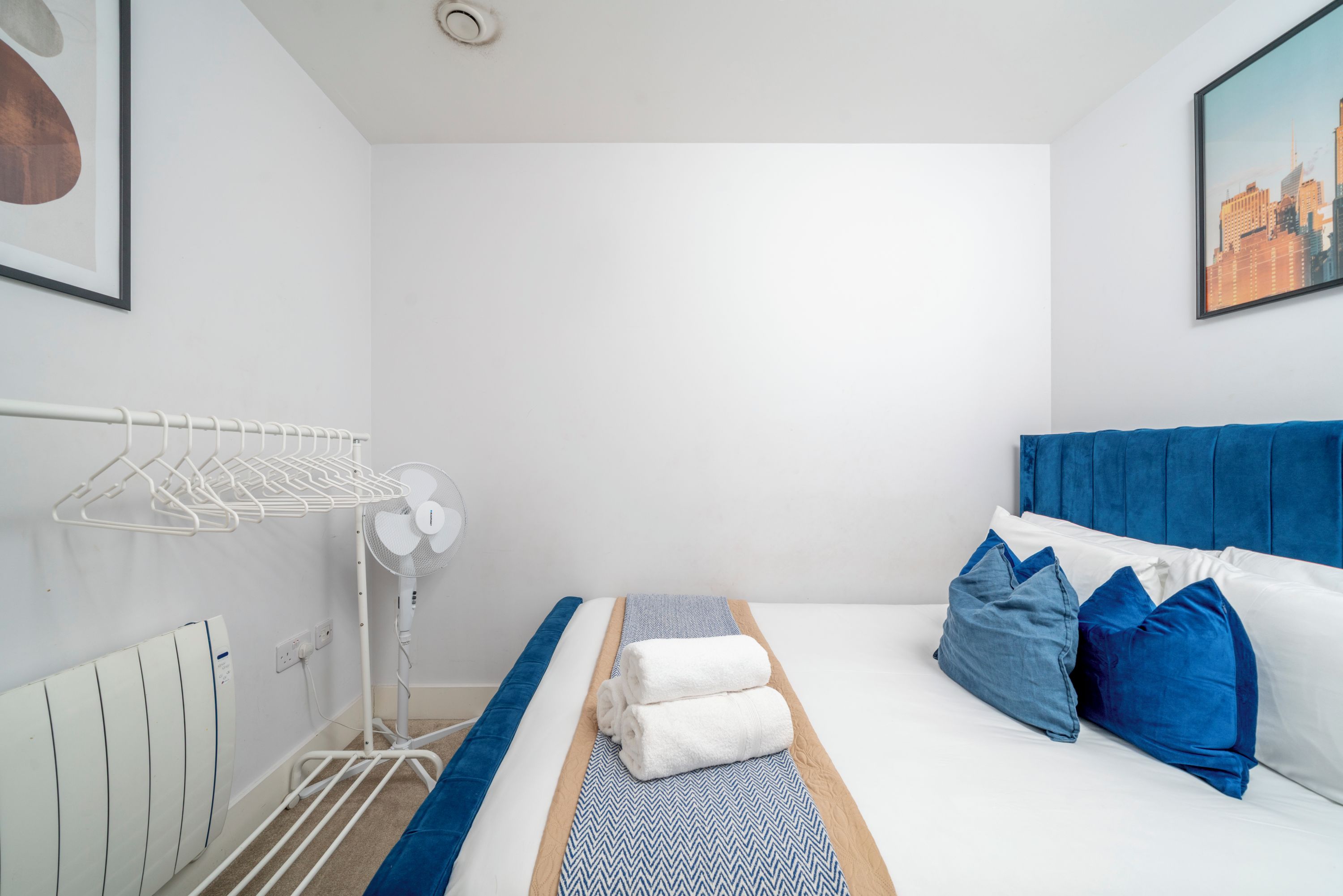 Une chambre d’hôtel avec un lit, un radiateur, un ventilateur et un tableau représentant un paysage urbain.