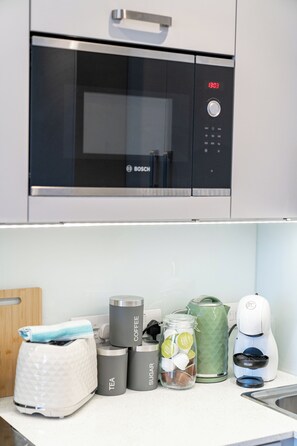 Una cocina con microondas Bosch, cafetera, hervidor de agua, un tarro de azúcar, un frasco de vidrio con bolsitas de té y una tetera verde.