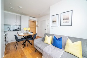 Un espace de vie moderne avec un canapé gris, des coussins noirs et jaunes, une table à manger en bois avec des chaises, et un coin cuisine doté de meubles blancs.