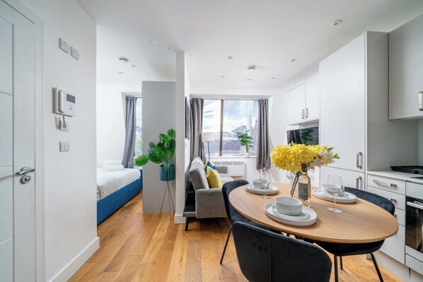 Une chambre d’hôtel moderne comprenant un lit, un coin repas avec une table et des chaises, une cuisine et une grande fenêtre donnant sur un paysage agréable.