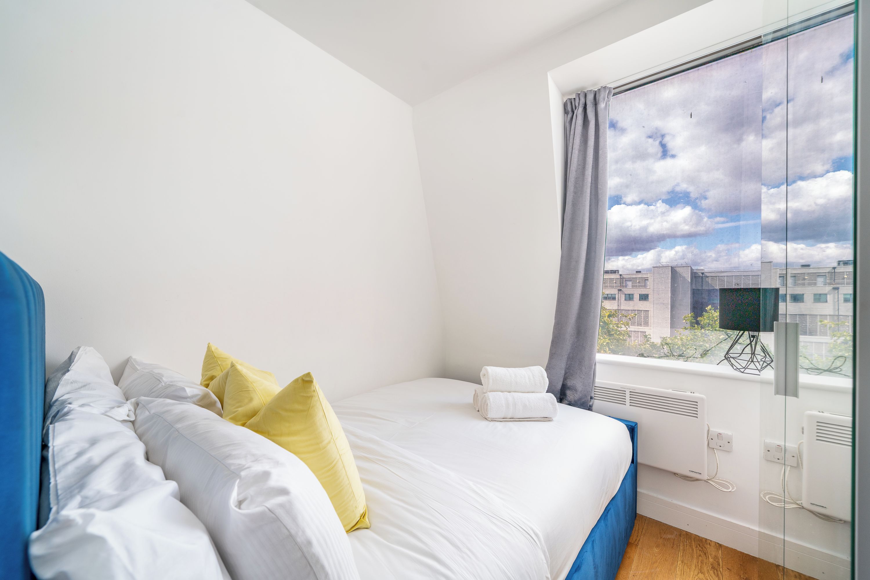 A hotel room with a bed, pillows, a window with a city view, and a thermostat.