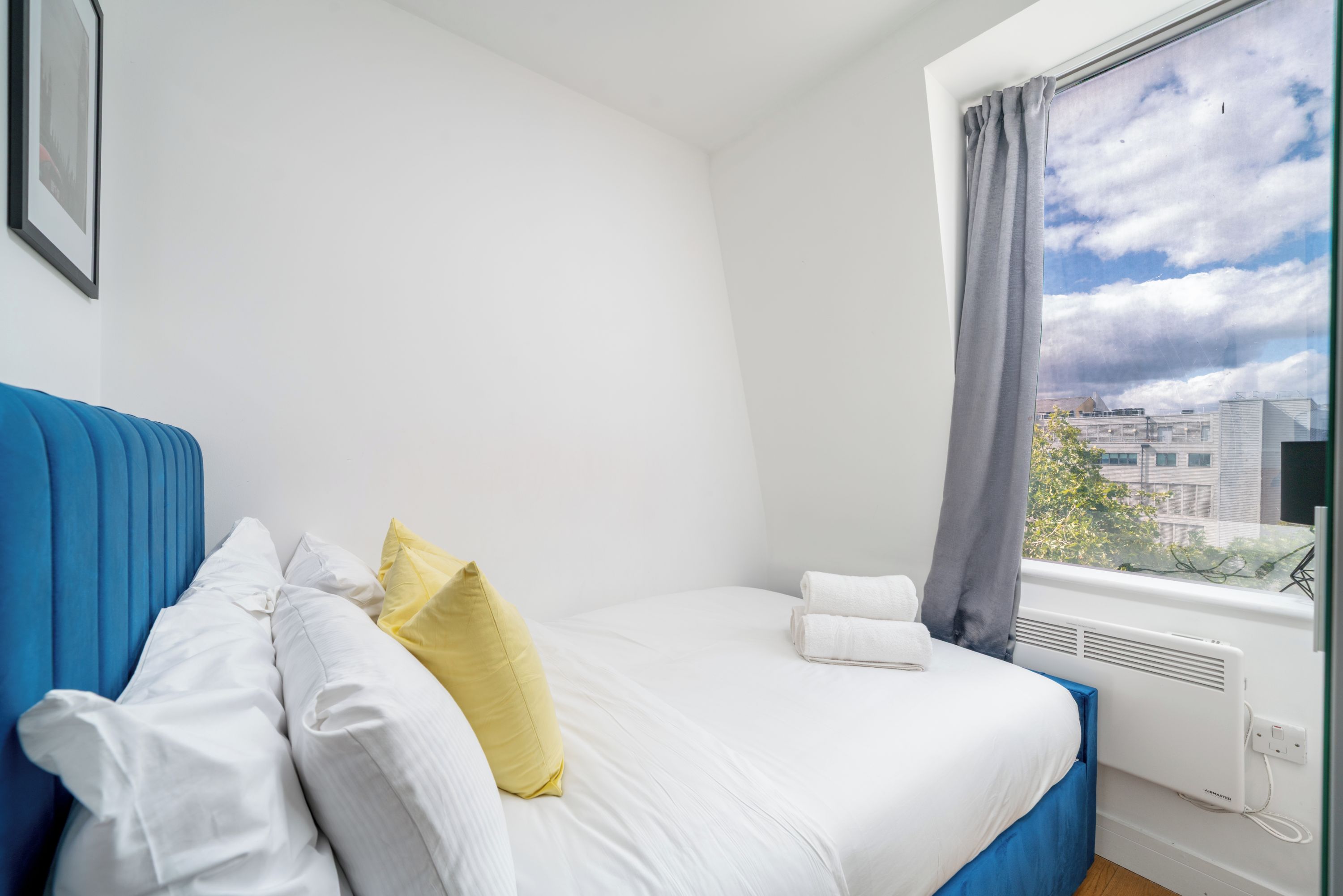Une chambre à coucher avec un lit, des oreillers, une fenêtre avec des rideaux et une vue sur des immeubles.