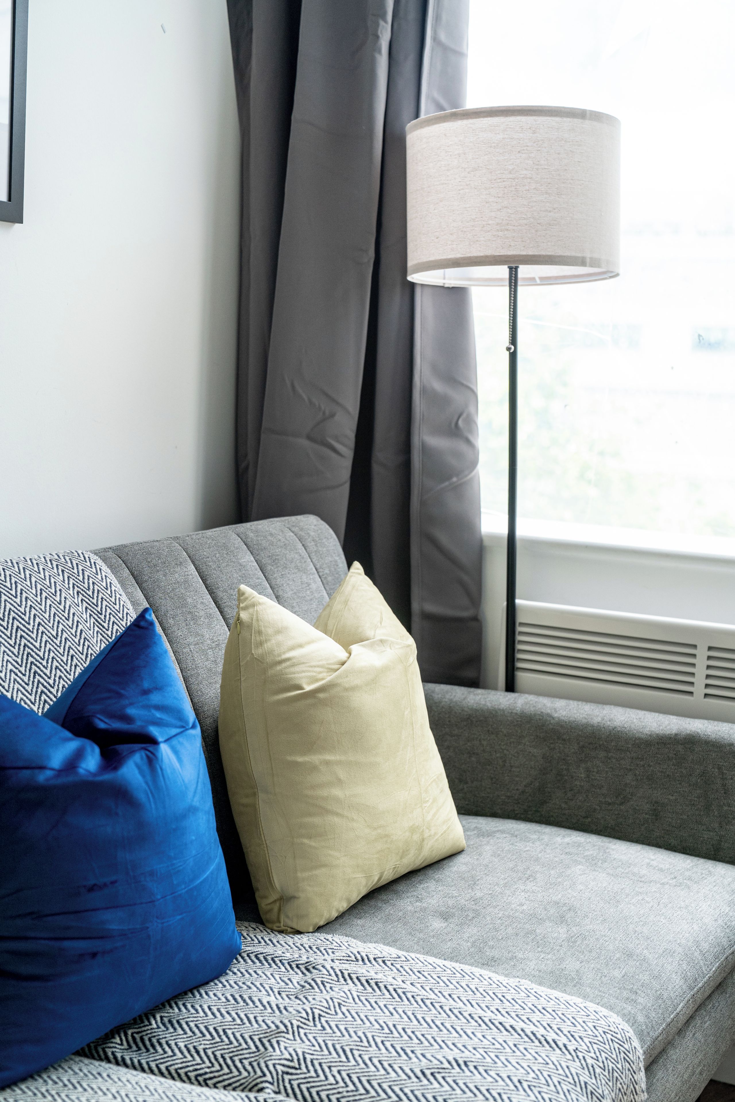 A grey sofa with a patterned throw, a blue pillow, and a beige pillow. A floor lamp is positioned to the right.