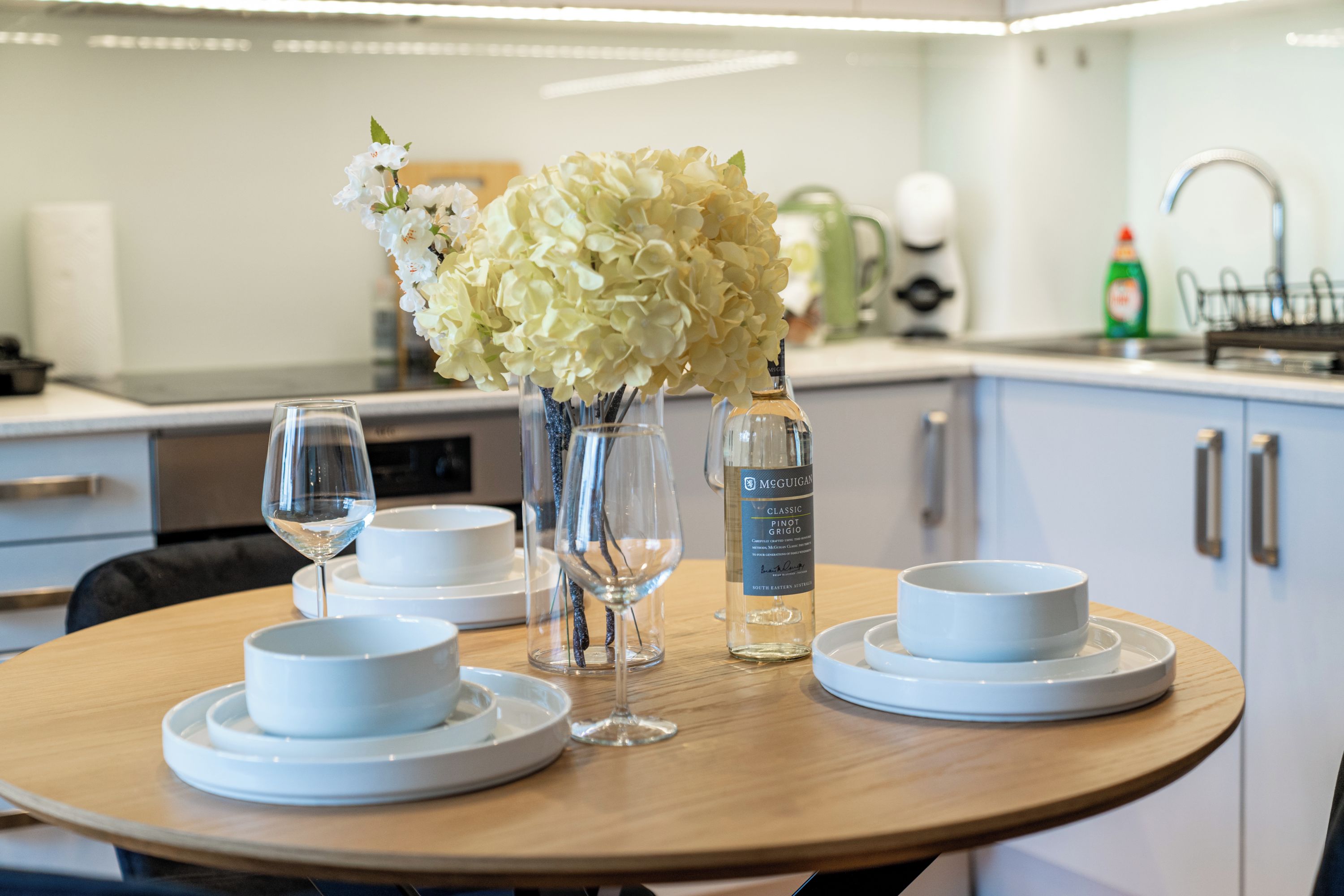 A round dining table with white plates, two wine glasses, and a bottle of McGuigan Classic Pinot Grigio.