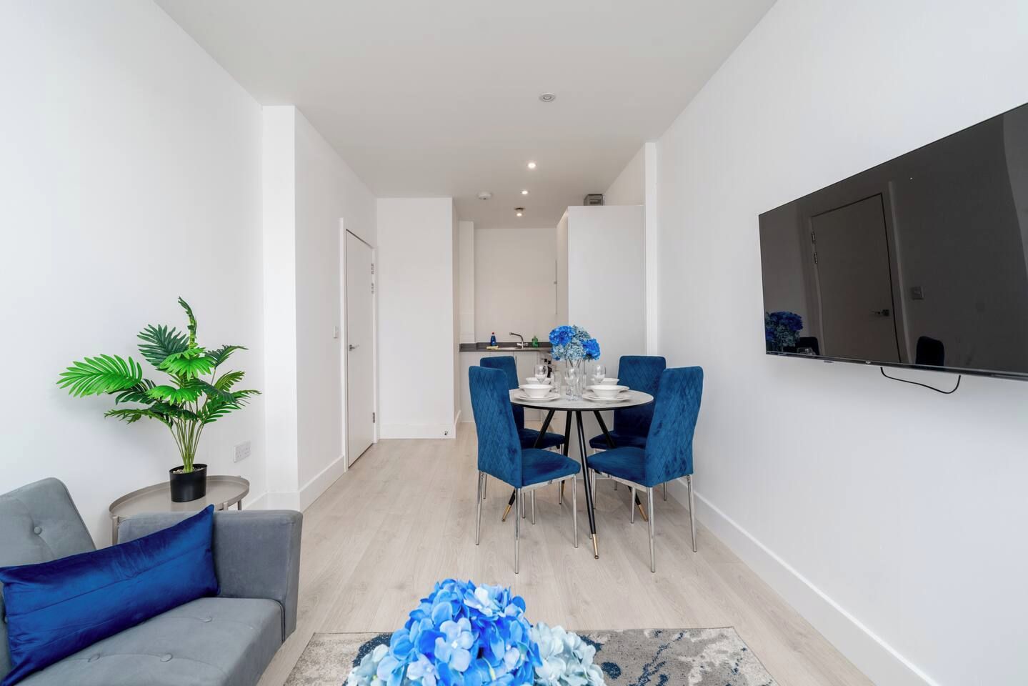 A modern living room with a grey sofa, a blue accent pillow, a round coffee table with a blue floral arrangement, and a dining area with blue chairs and a white table.