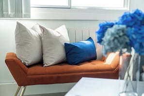 Ein Sofa mit drei Kissen, eine Vase mit Blumen und ein Fenster im Hintergrund.