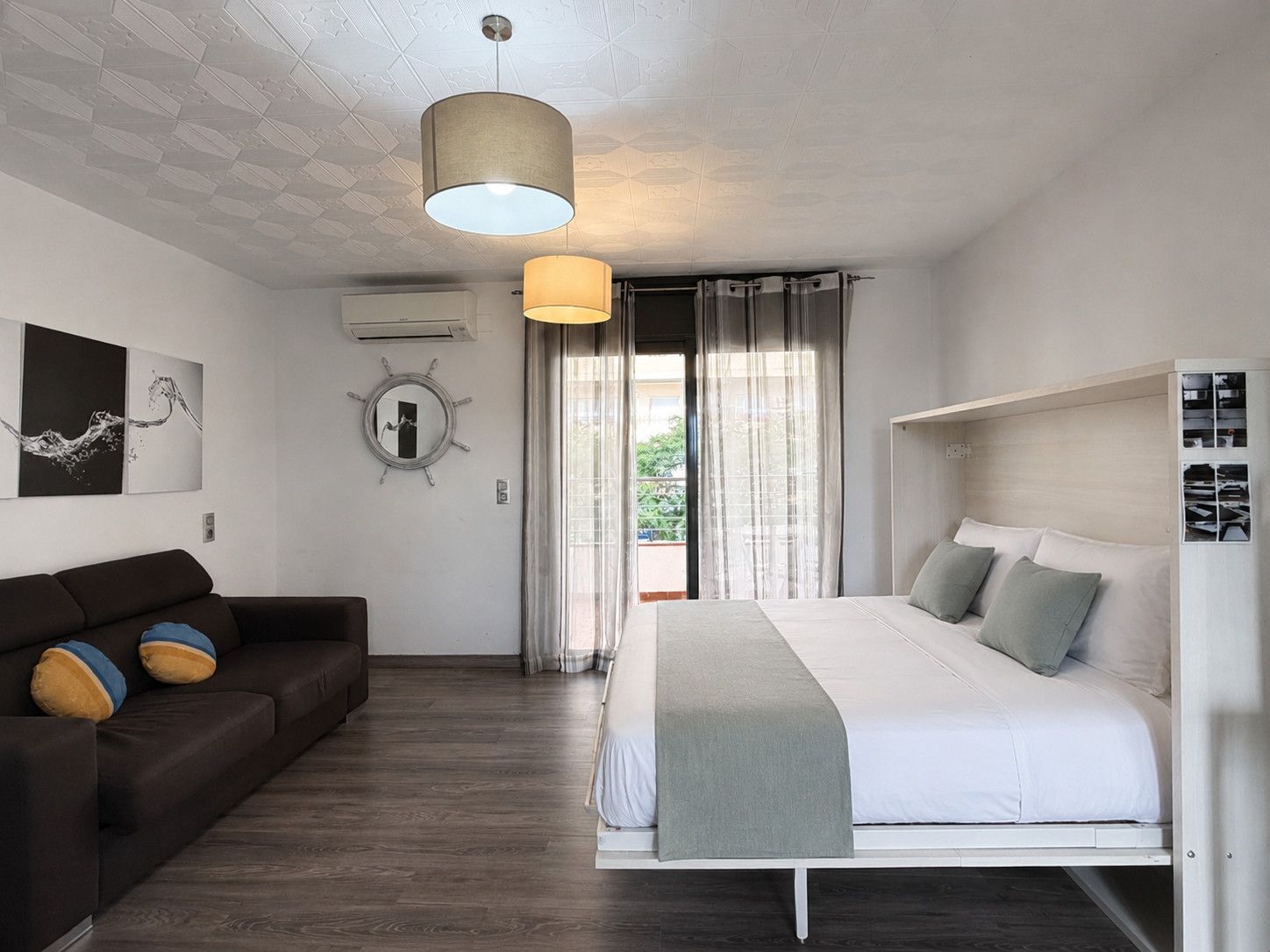 Un dormitorio moderno con cama, sofá, lámpara colgante, ventana con cortinas y un equipo de aire acondicionado instalado en la pared.
