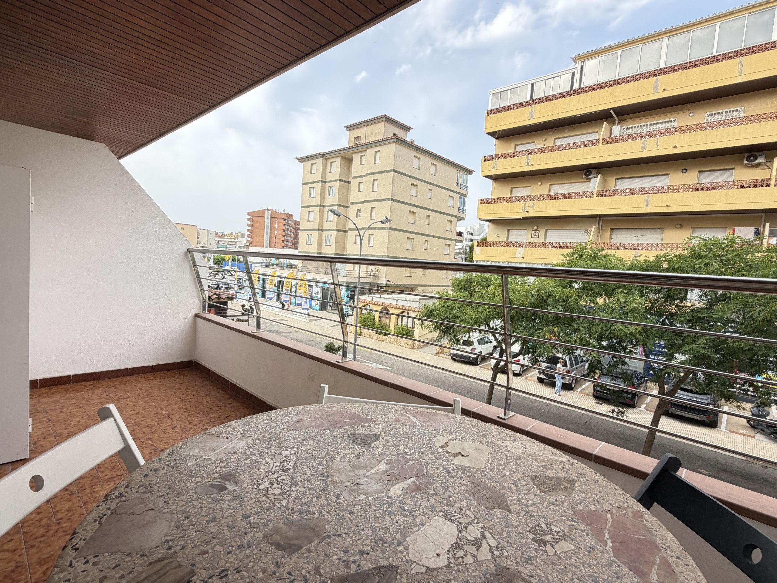Un balcón con una mesa redonda y sillas, con vista a una calle con coches estacionados y edificios.