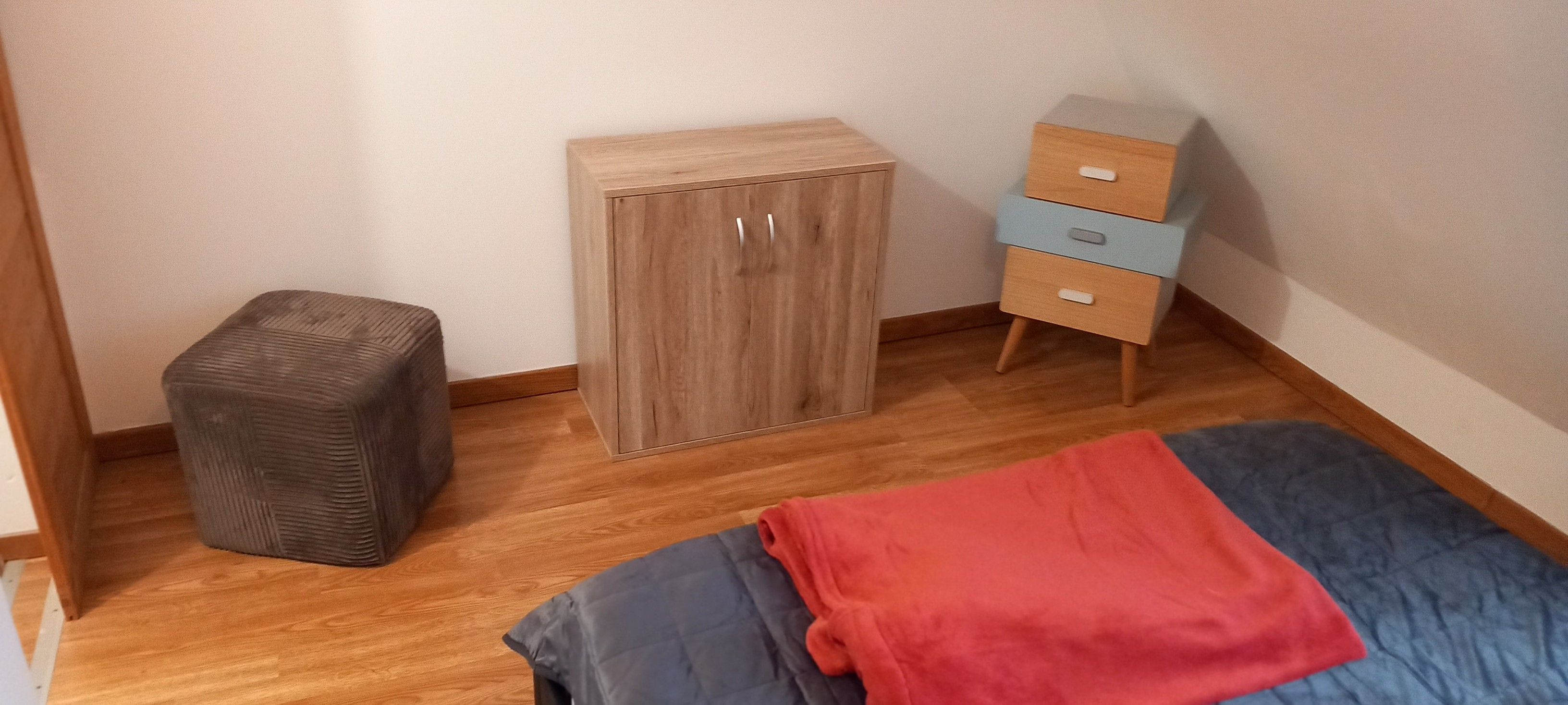 Une pièce comprenant une armoire en bois, une petite commode, un pouf gris et un lit recouvert d’une couverture bleue et rouge.