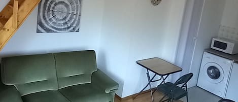 Una sala de estar con un sofá verde, una mesita, una silla, una escalera de madera y una lavadora.
