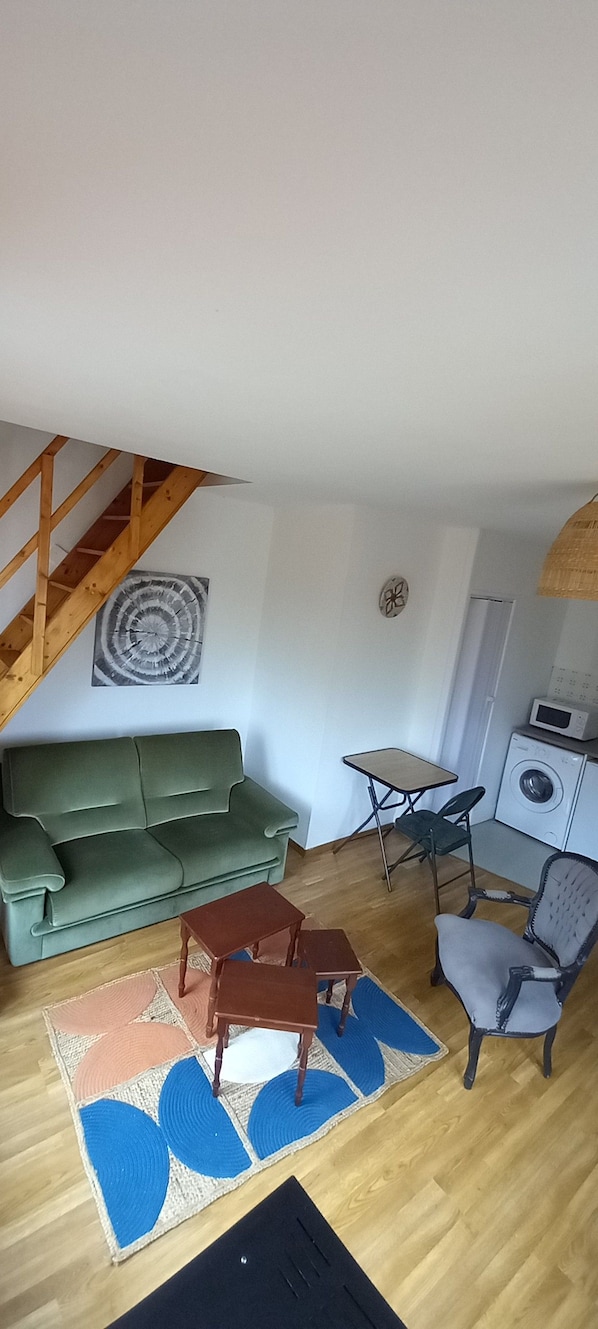 A living room with a green sofa, a small table, a chair, a wooden staircase, and a washing machine. - Apartment 'L'étoile De L'échappée En Thiérache' with Wi-Fi (Martigny)