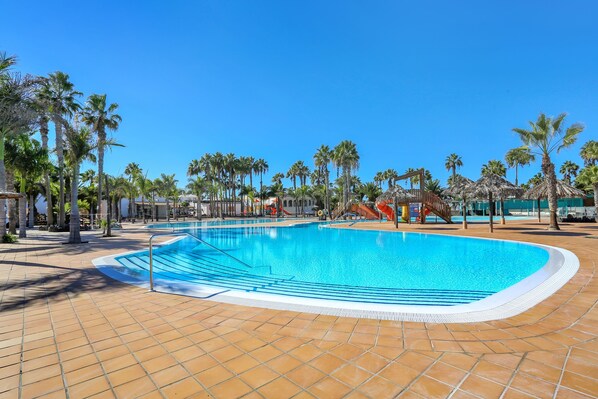 Una piscina con solárium de baldosas, palmeras y un tobogán al fondo.