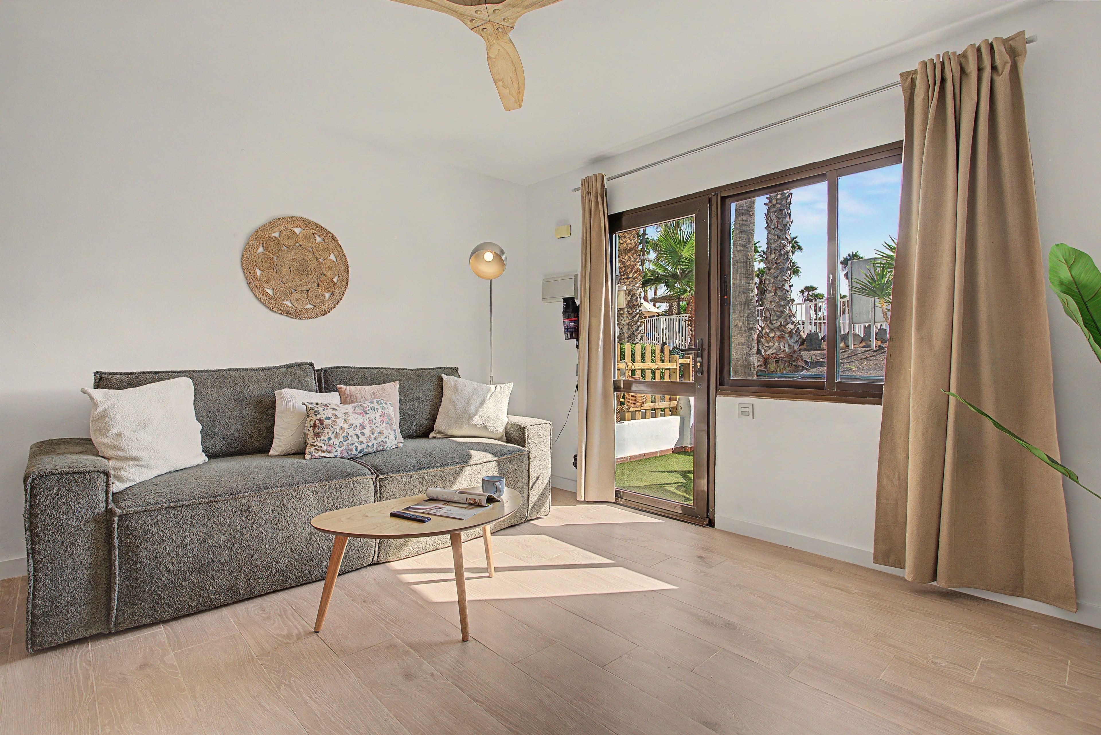 Una sala de estar con un sofá gris, una mesa de centro y un amplio ventanal corredizo que da a un espacio exterior con plantas.